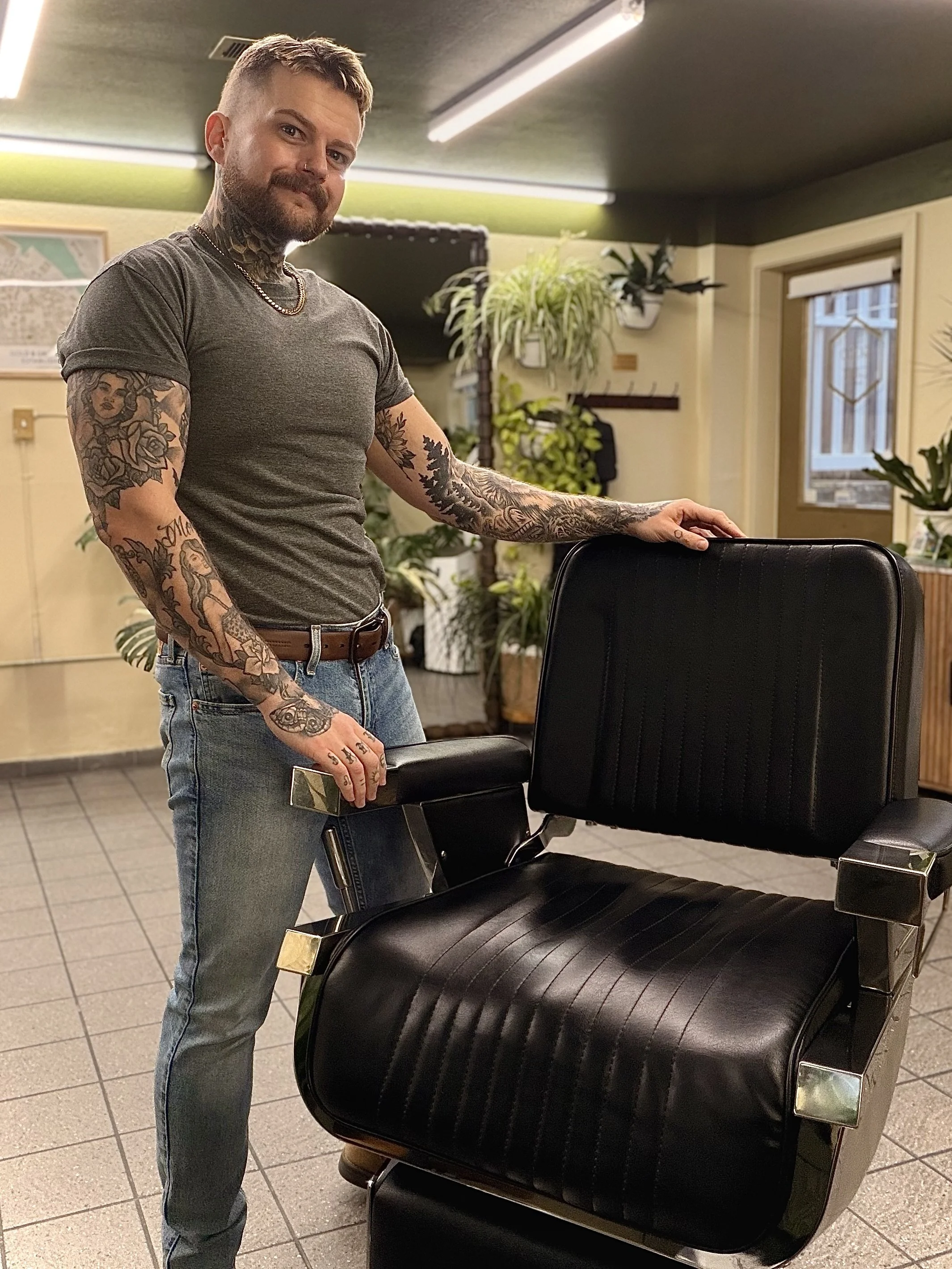 A tattooed man with short hair and a beard, wearing a grey T-shirt and jeans, standing in a room with plants and a window, holding a phone in his right hand, with his left hand on a black office chair.