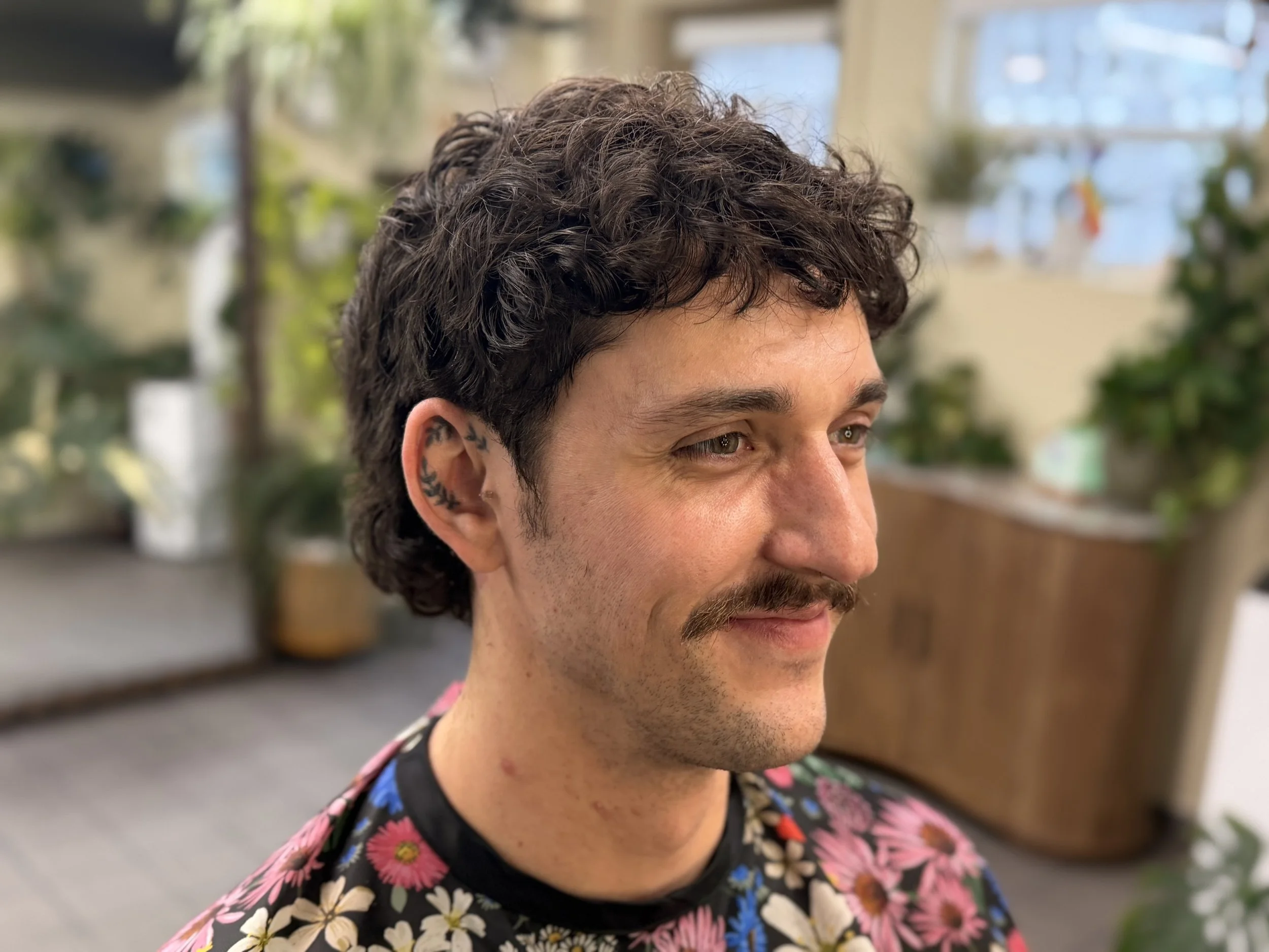 Close-up of a man with dark, curly hair and a tattoo behind his ear, wearing a floral shirt, indoors with blurred background.