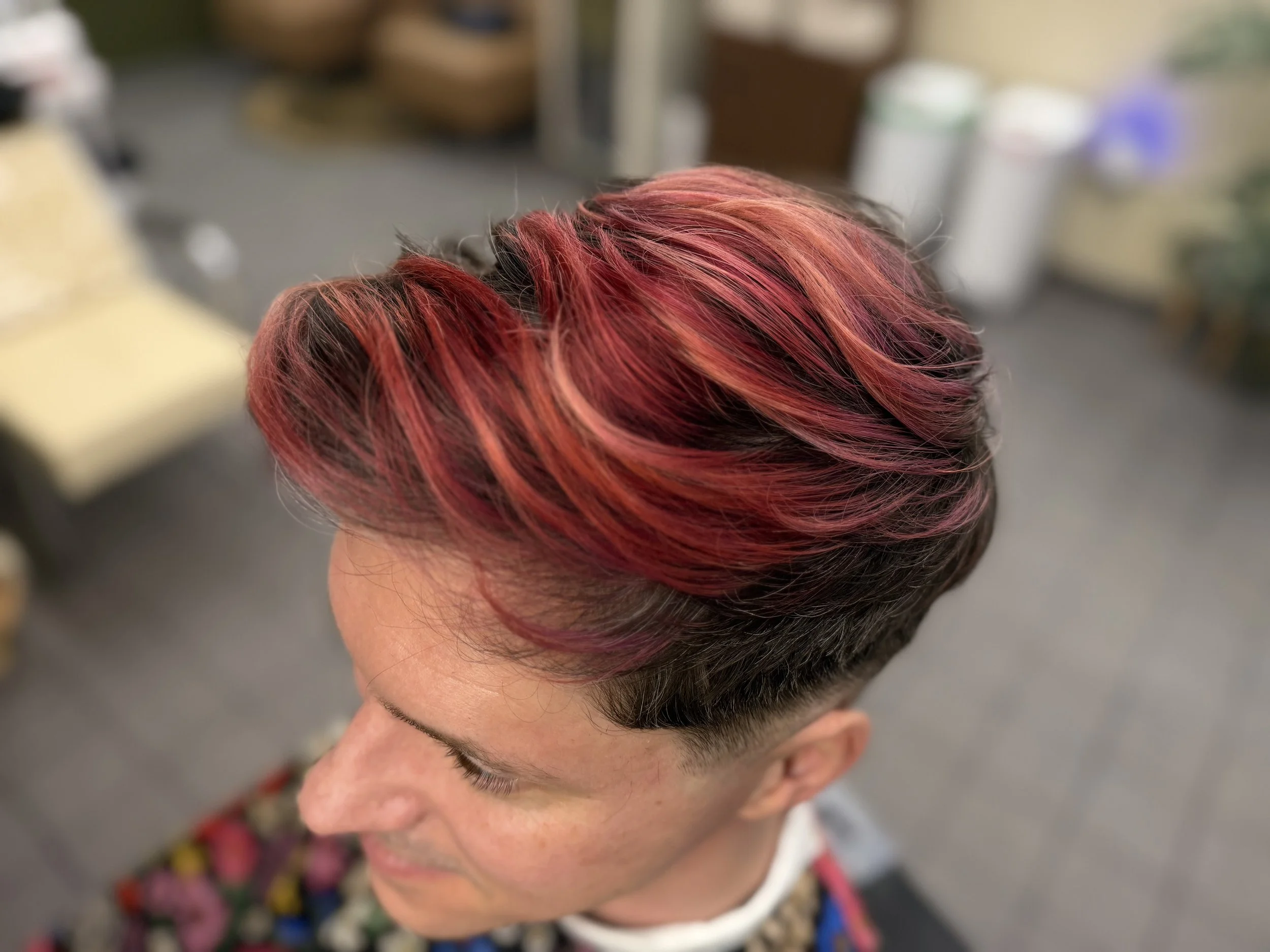A person with short hair dyed in shades of red, pink, and orange, styled with soft waves, in an indoor setting with a blurred background.