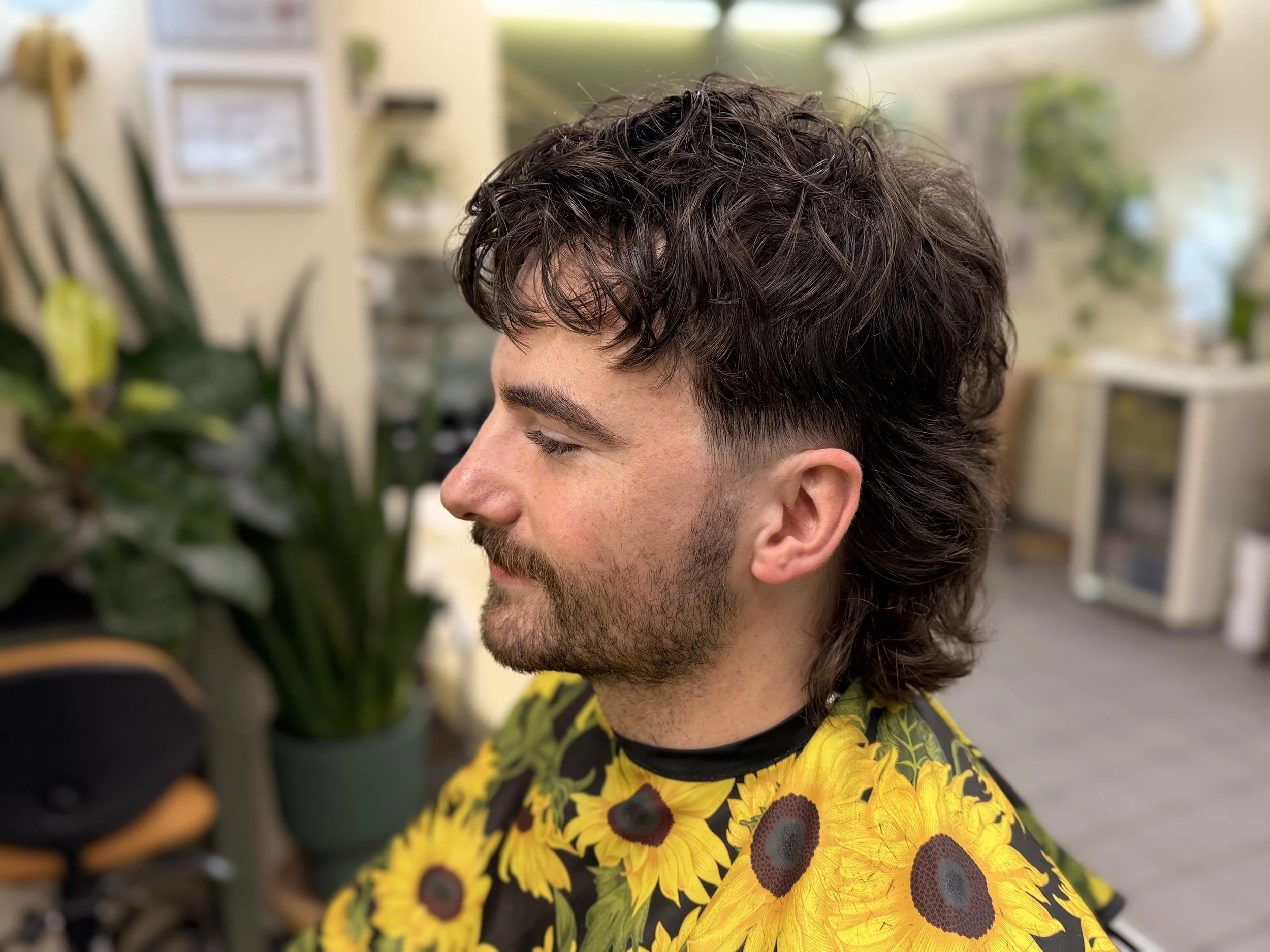 A young man with dark curly hair and a beard, smiling gently with eyes closed, wearing a vibrant shirt with sunflower patterns, standing in a room with plants and pictures on the wall.