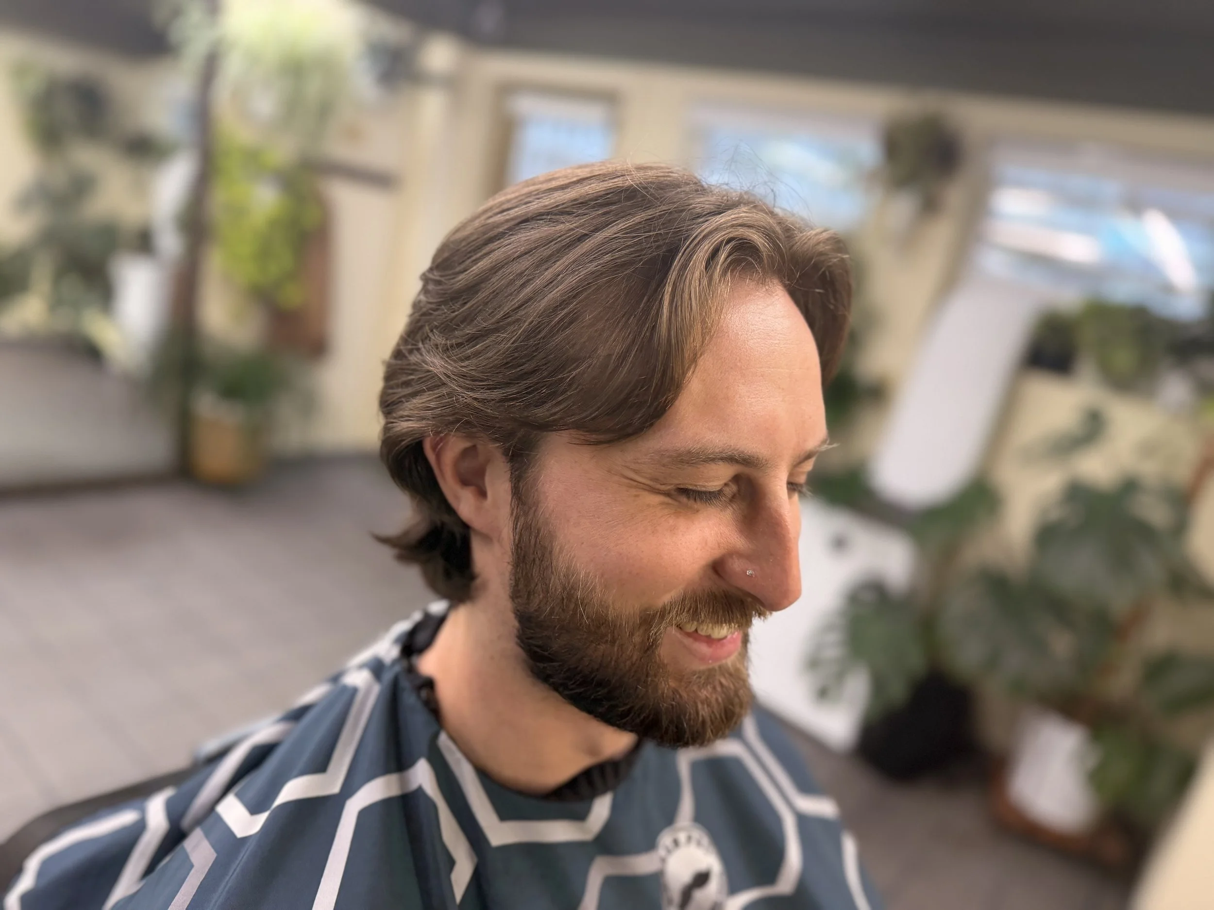 Close-up of a smiling man with brown hair and a beard, wearing a blue and white striped shirt, outdoors near a window and plant.