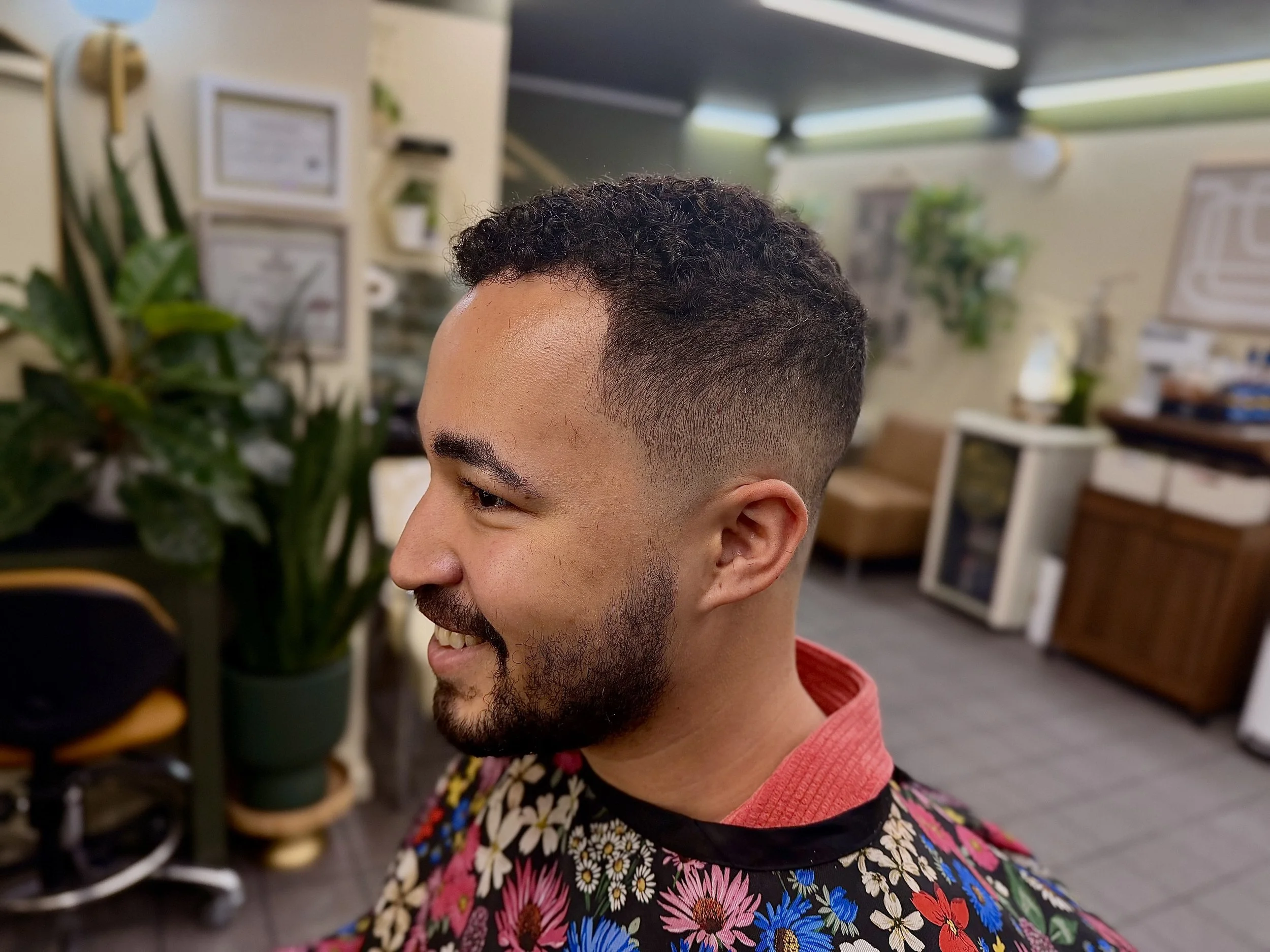 A man with short, curly hair and a beard smiling while looking down. He wears a shirt with a colorful floral pattern and a red collar. The background shows a plant and some office or shop furnishings.