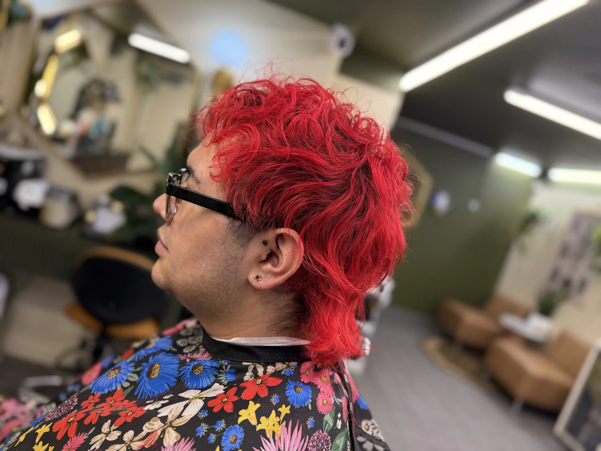 Profile view of woman with vibrant red, curly hair; wearing glasses and floral-patterned shirt; background of hair salon interior.