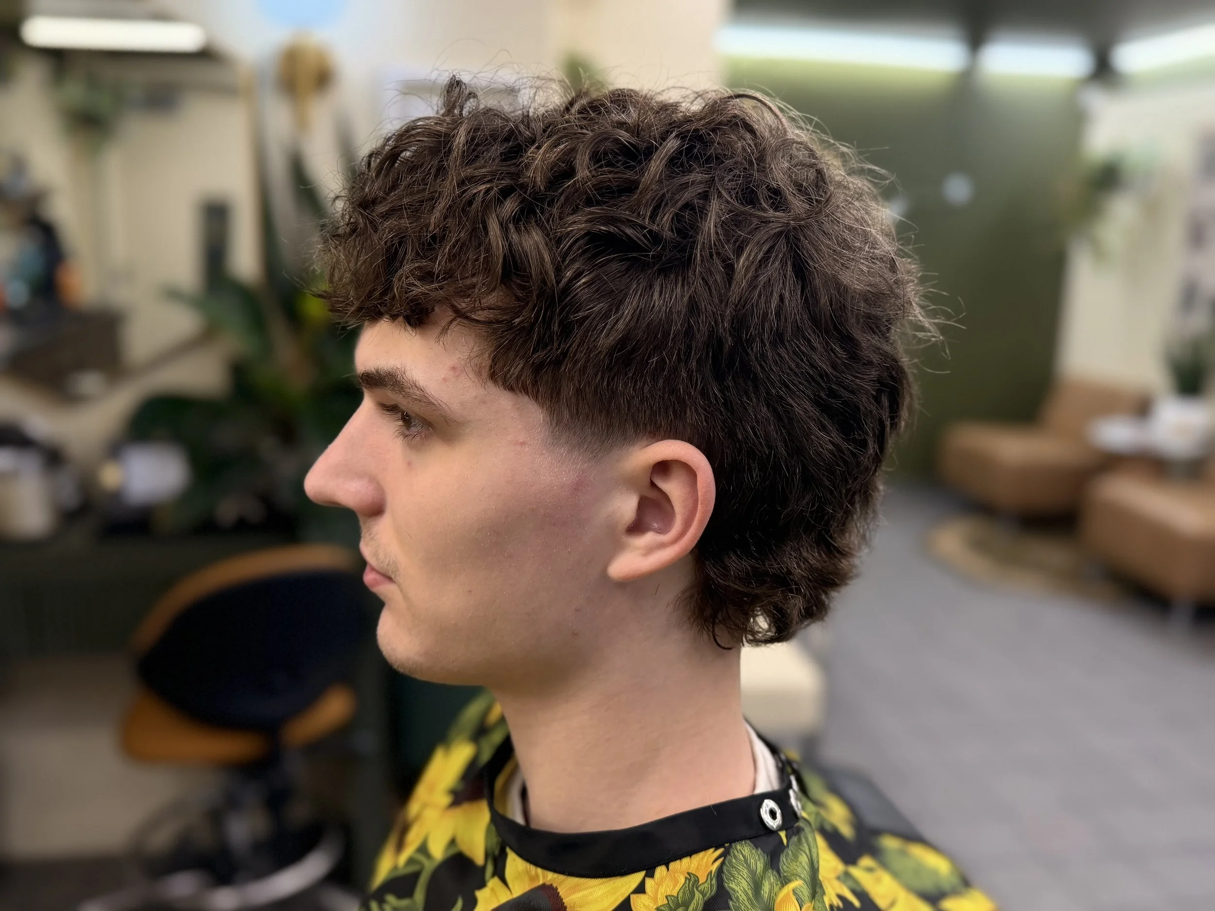 Side profile of a young man with curly brown hair, wearing a black shirt with a sunflower pattern, in a barbershop or hair salon.