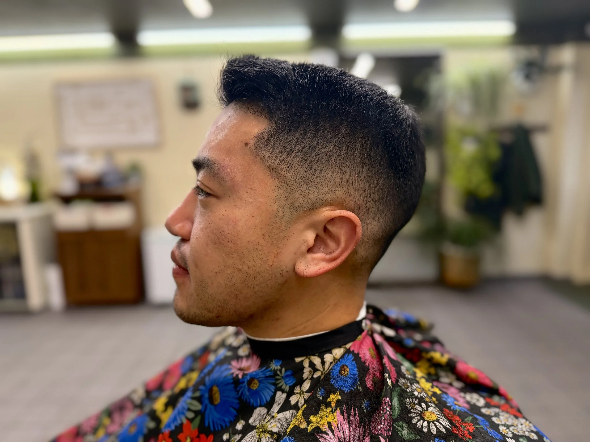 Close-up of a young man's head with a textured haircut, wearing a floral cape in a barbershop or salon.