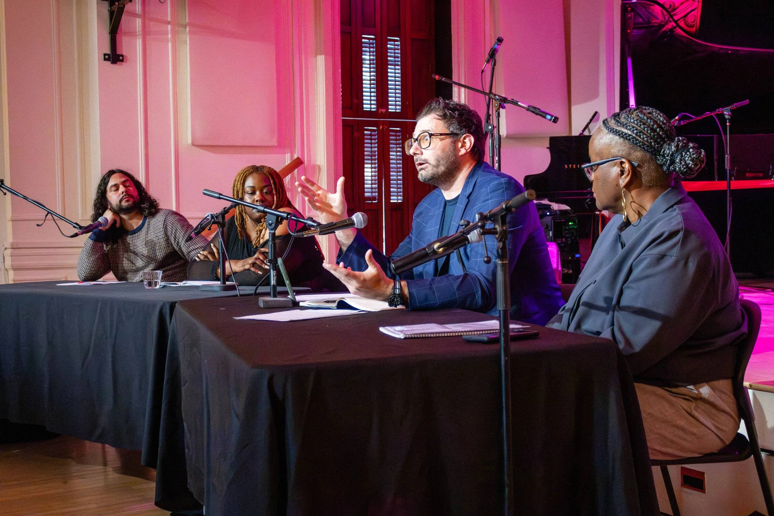 A panel discussion with four people seated at a table in a room with wooden shutters and a grand piano in the background. The speaker in the center is speaking and gesturing with his hands.