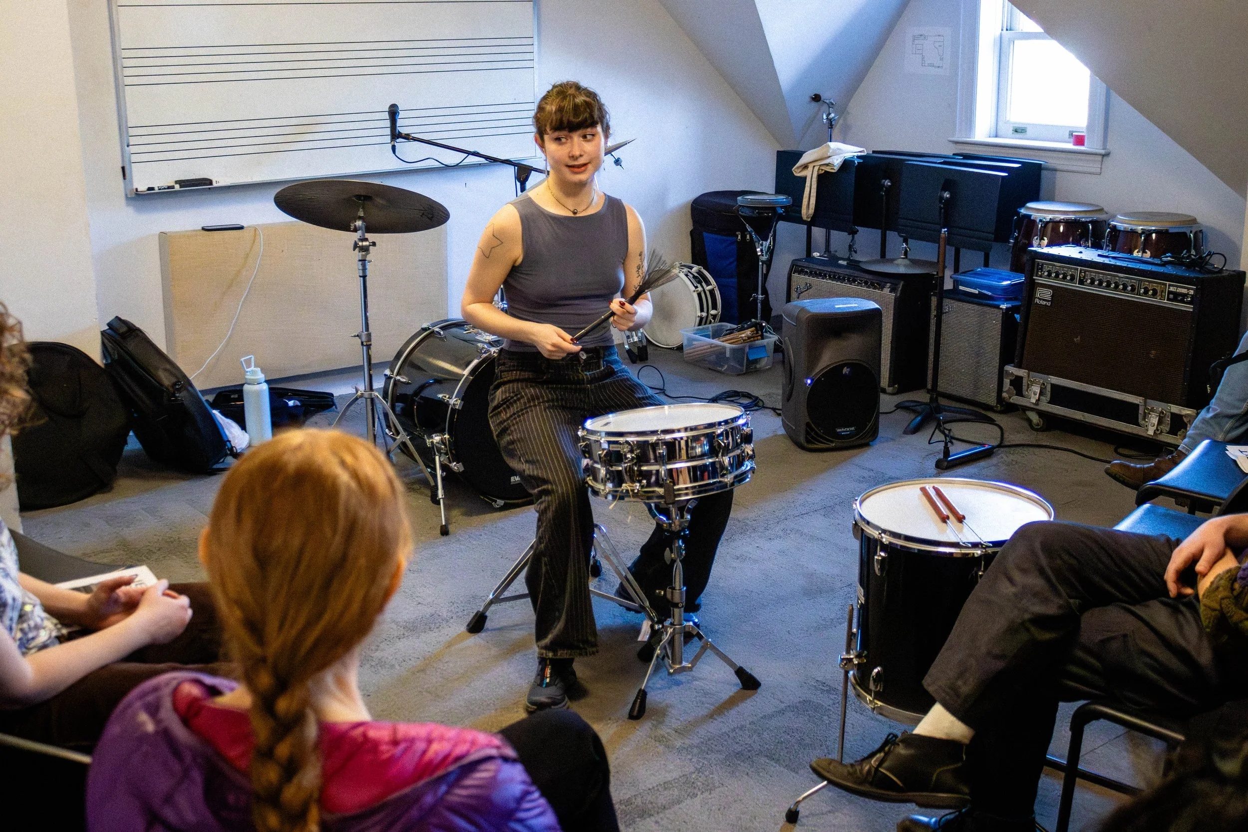 A young woman in a sleeveless gray top and striped pants appears to be playing drums in a music classroom. She is surrounded by musical equipment, including drums, amplifiers, and a whiteboard with musical staff lines. Several children are sitting an