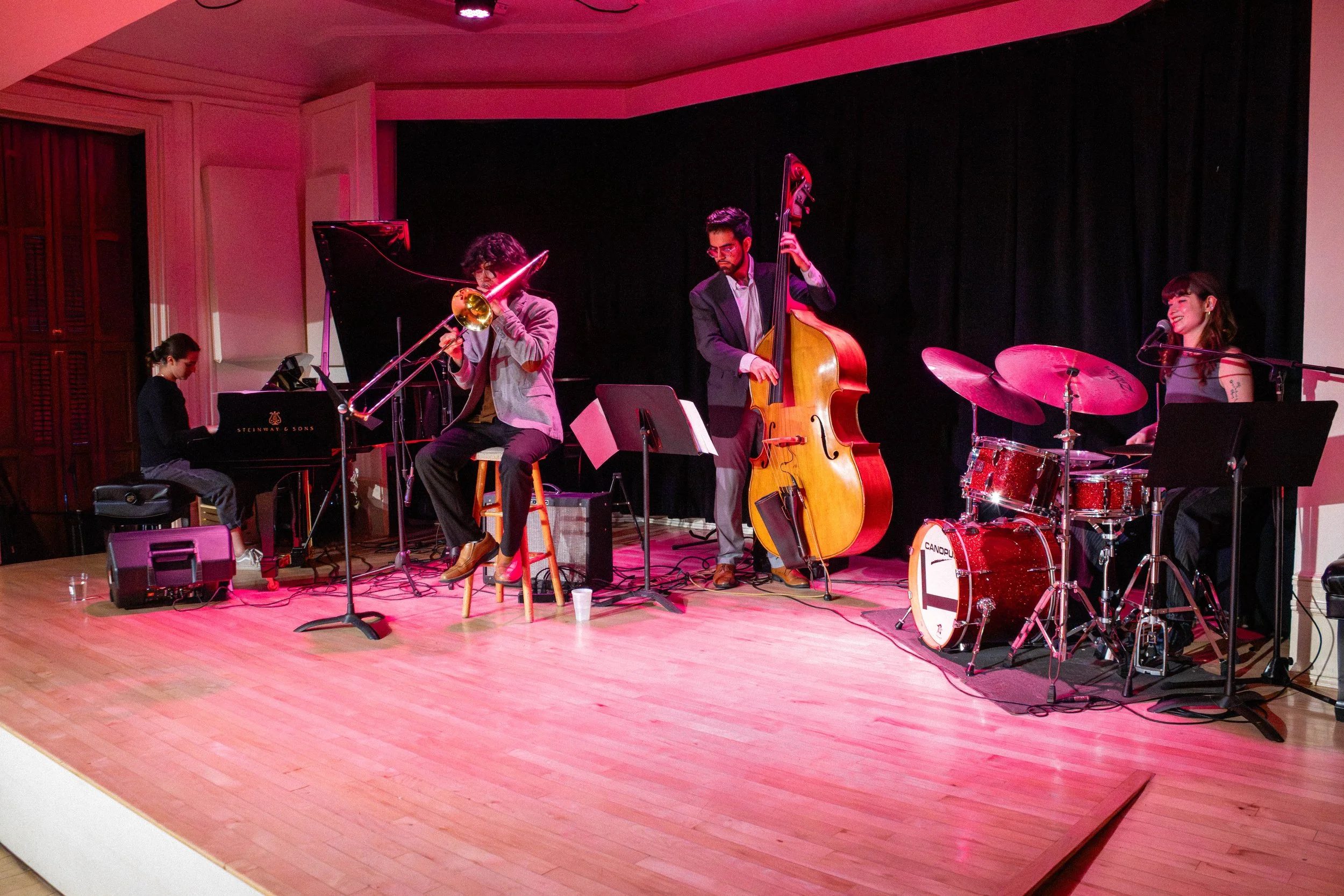 A jazz band performing on stage with a piano, trombone, double bass, and drum set, lit with pink and purple stage lighting.