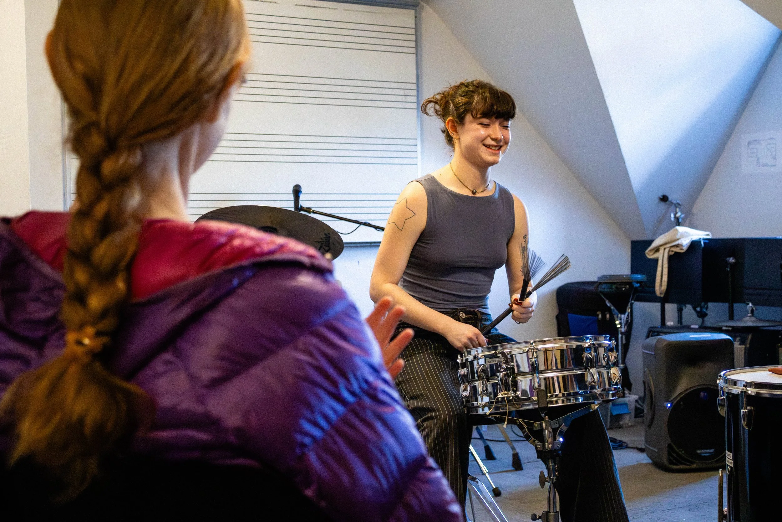 A young woman with tattoos on her arms smiling while playing percussion instruments, with another person with braided hair and a purple jacket in the foreground, in a music practice room with musical staff lines on the wall and musical equipment.