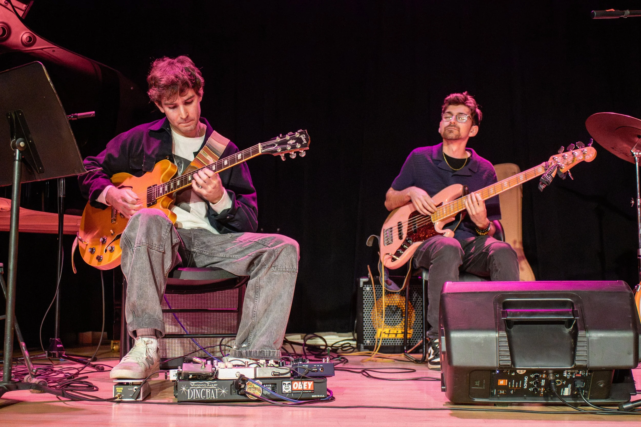 Two musicians seated on stage playing electric guitars, surrounded by audio equipment, with a dark background and stage lighting.