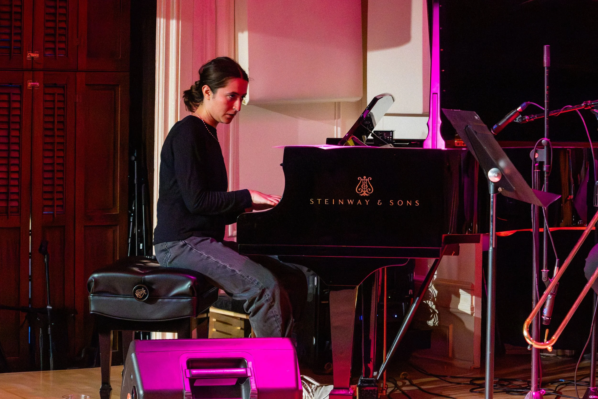 A woman playing a black Steinway & Sons grand piano on stage, illuminated by pink stage lighting, with microphone and music stand nearby.