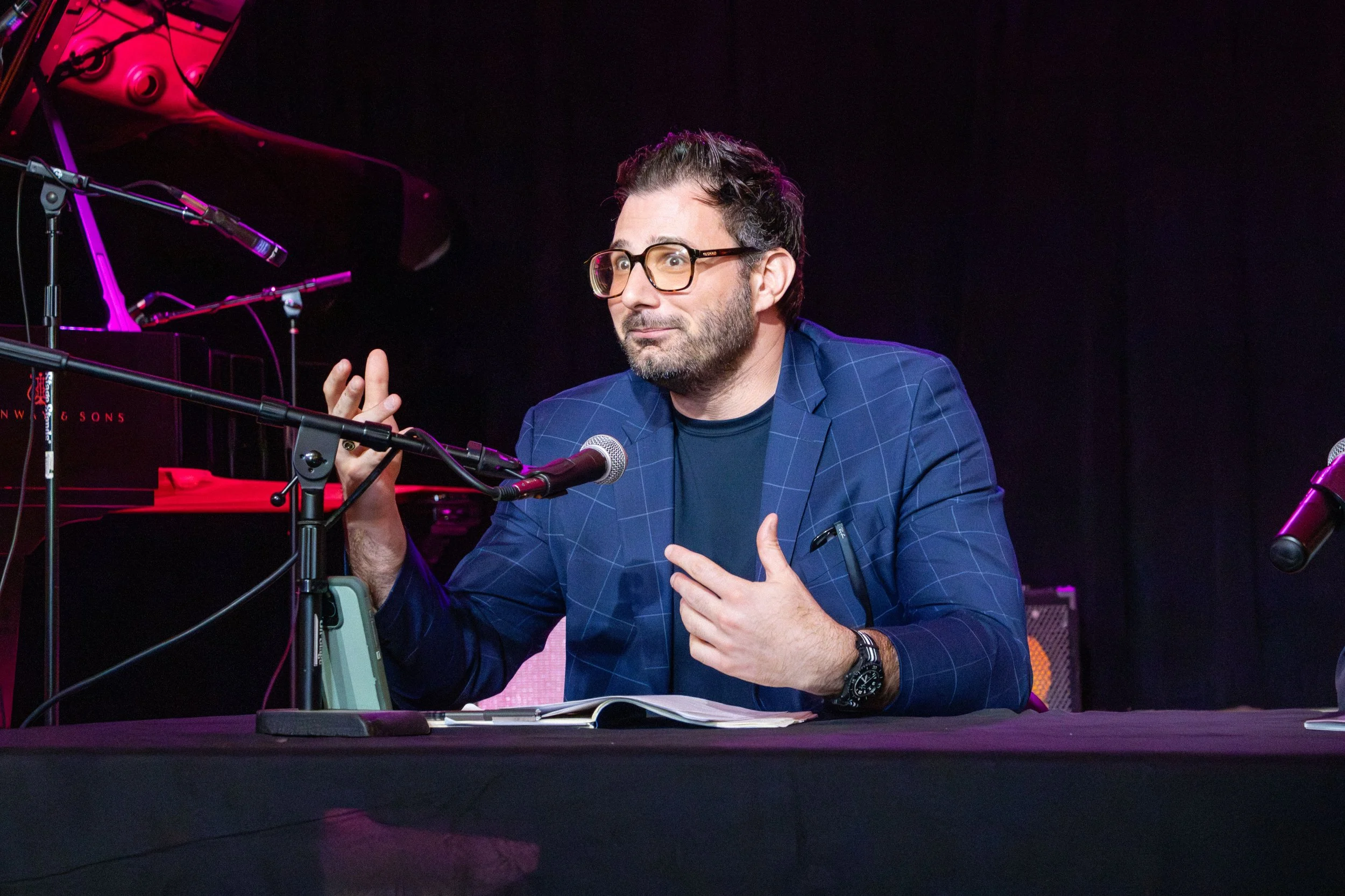 A man with glasses and a beard is seated at a table with a black tablecloth, speaking into a microphone. He is dressed in a dark checked blazer and a black T-shirt, with a notebook open in front of him. Behind him, there are microphones and audio equ