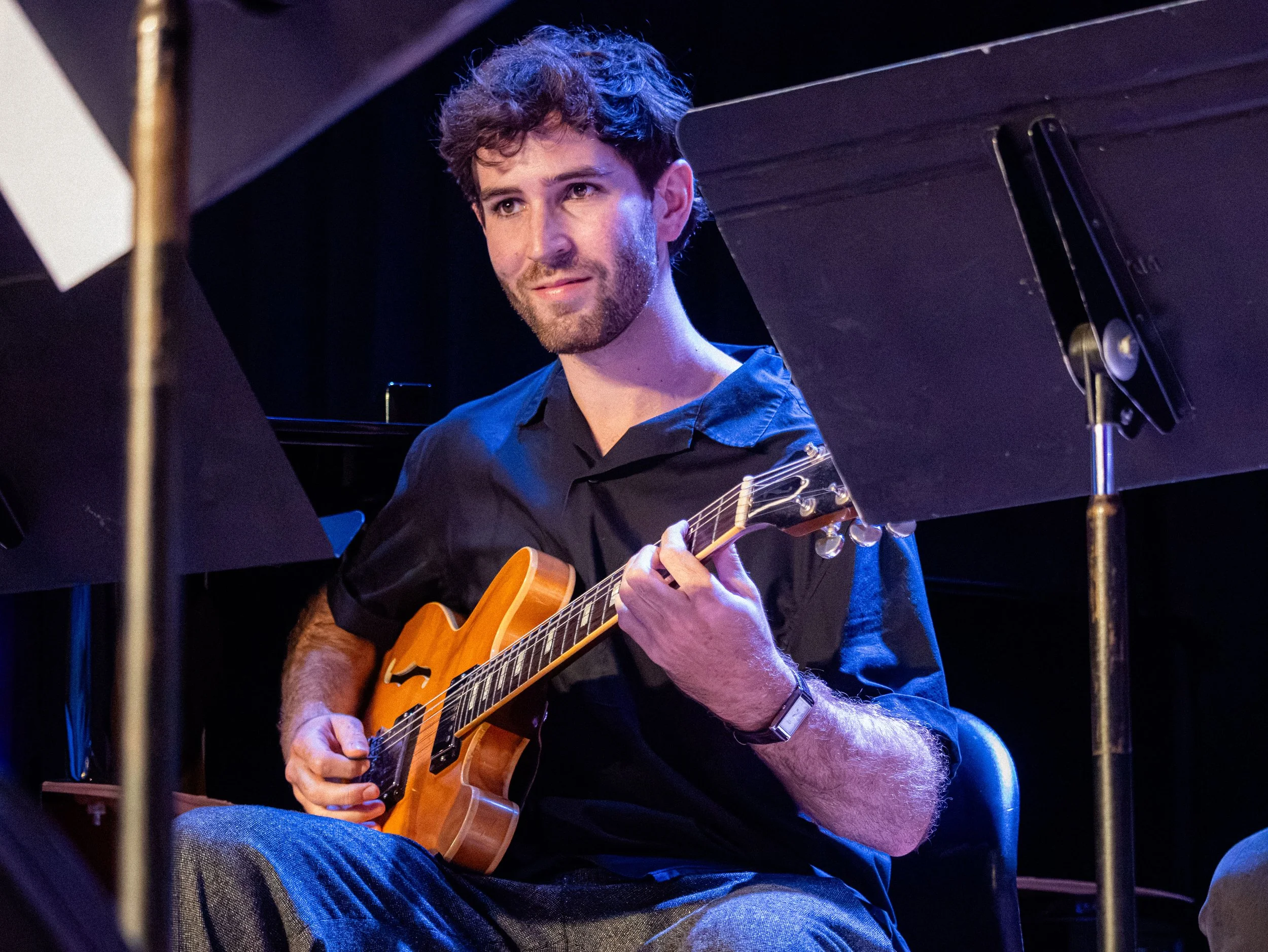 A man with curly hair and a beard playing an acoustic guitar on stage, surrounded by music stands and dark background.