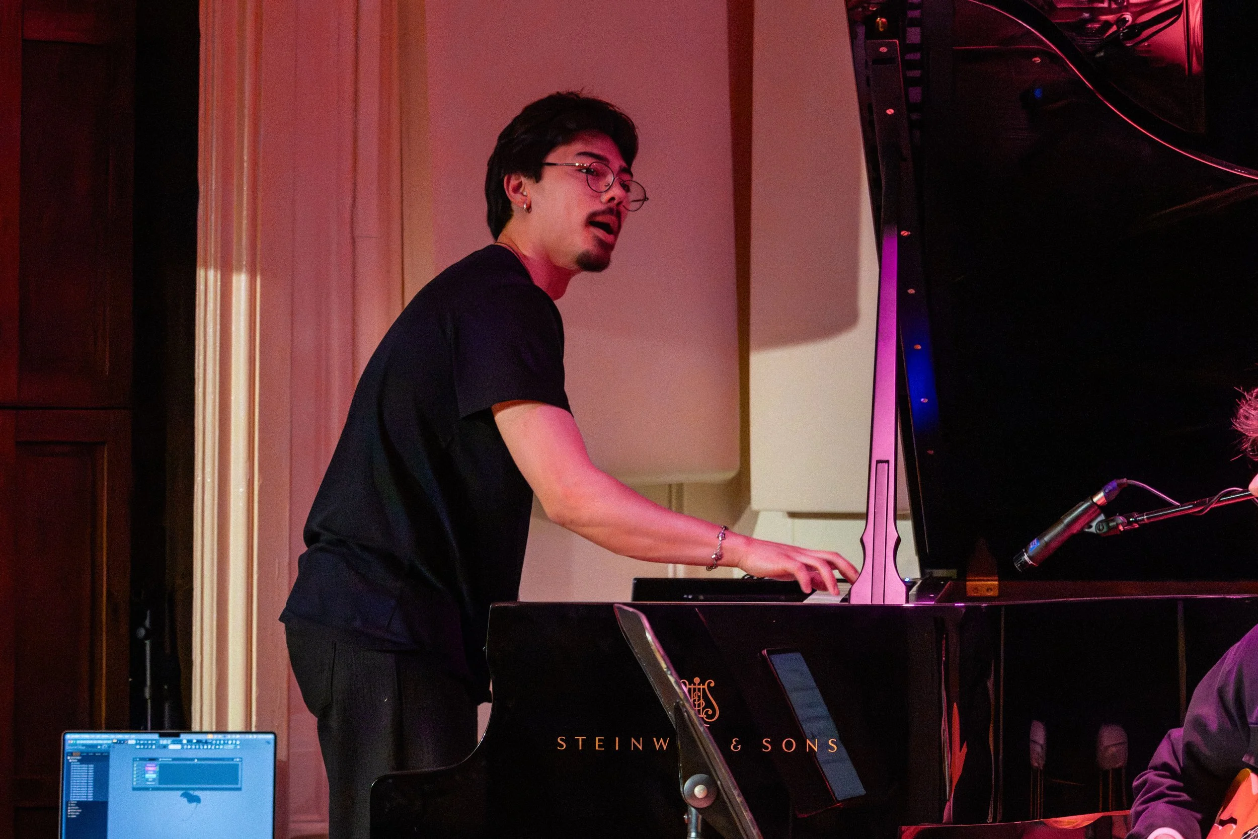A young man with glasses and dark hair playing a grand piano at a music performance.