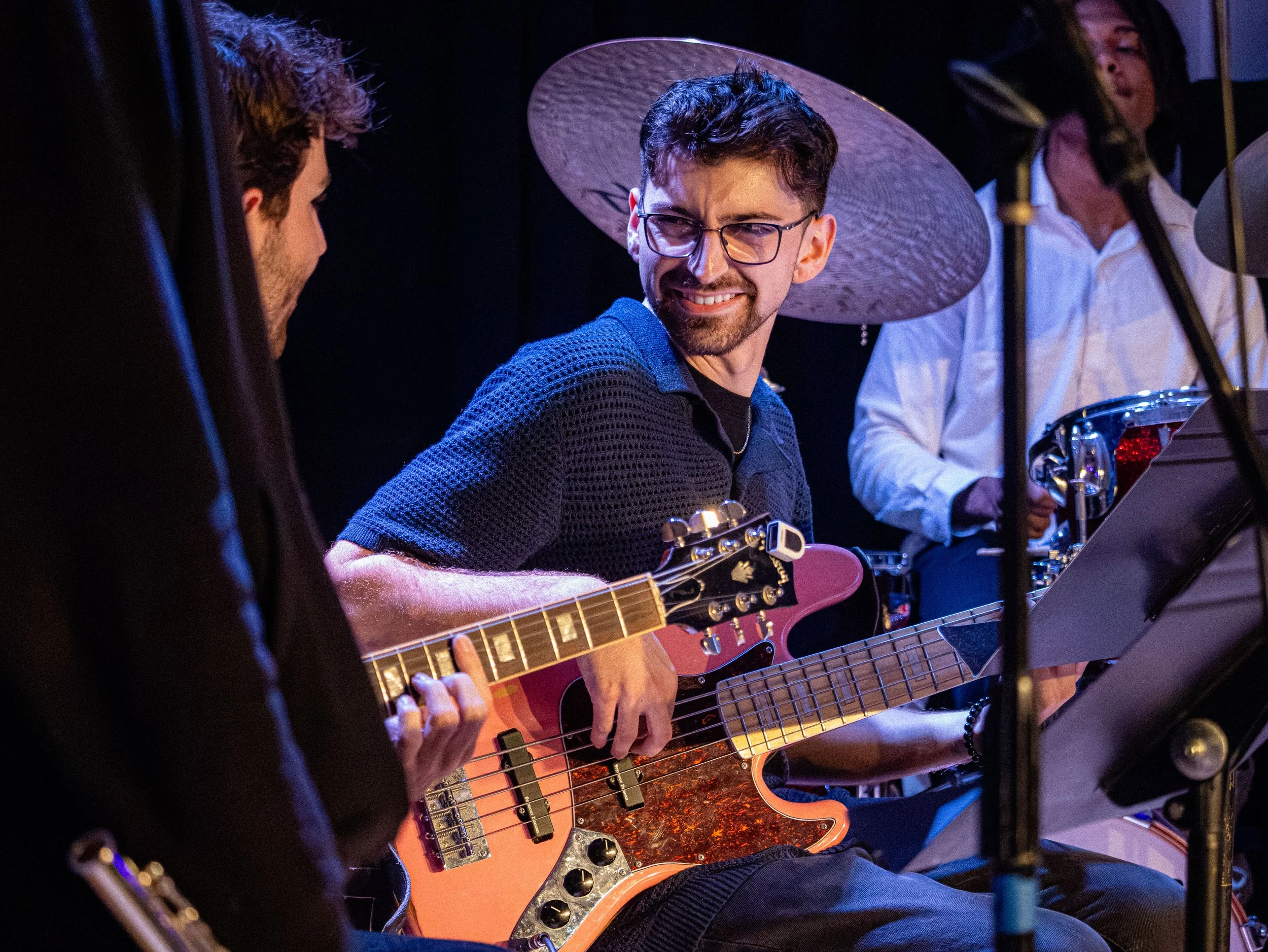 A group of people, including a young man with glasses, are playing guitars in a dimly lit setting, possibly during a band practice or performance.