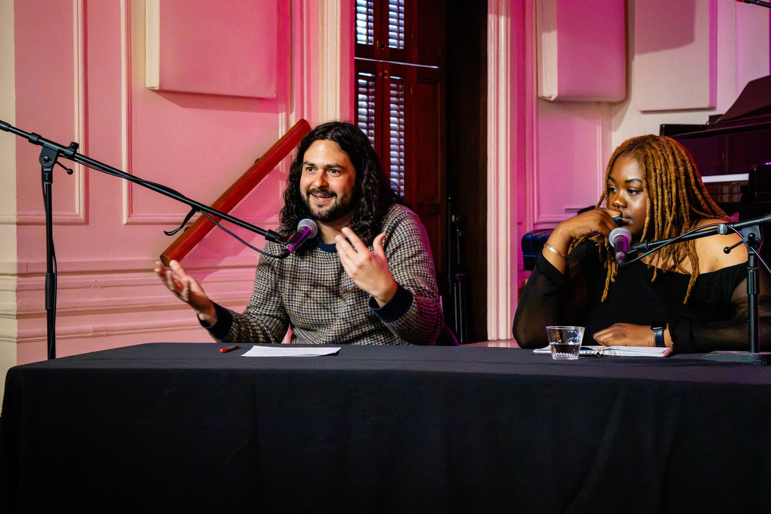 Two people sitting at a table with microphones, a man with long dark hair holding a pointer and speaking, a woman with long dreadlocks listening thoughtfully, in a decorated room with pink walls.