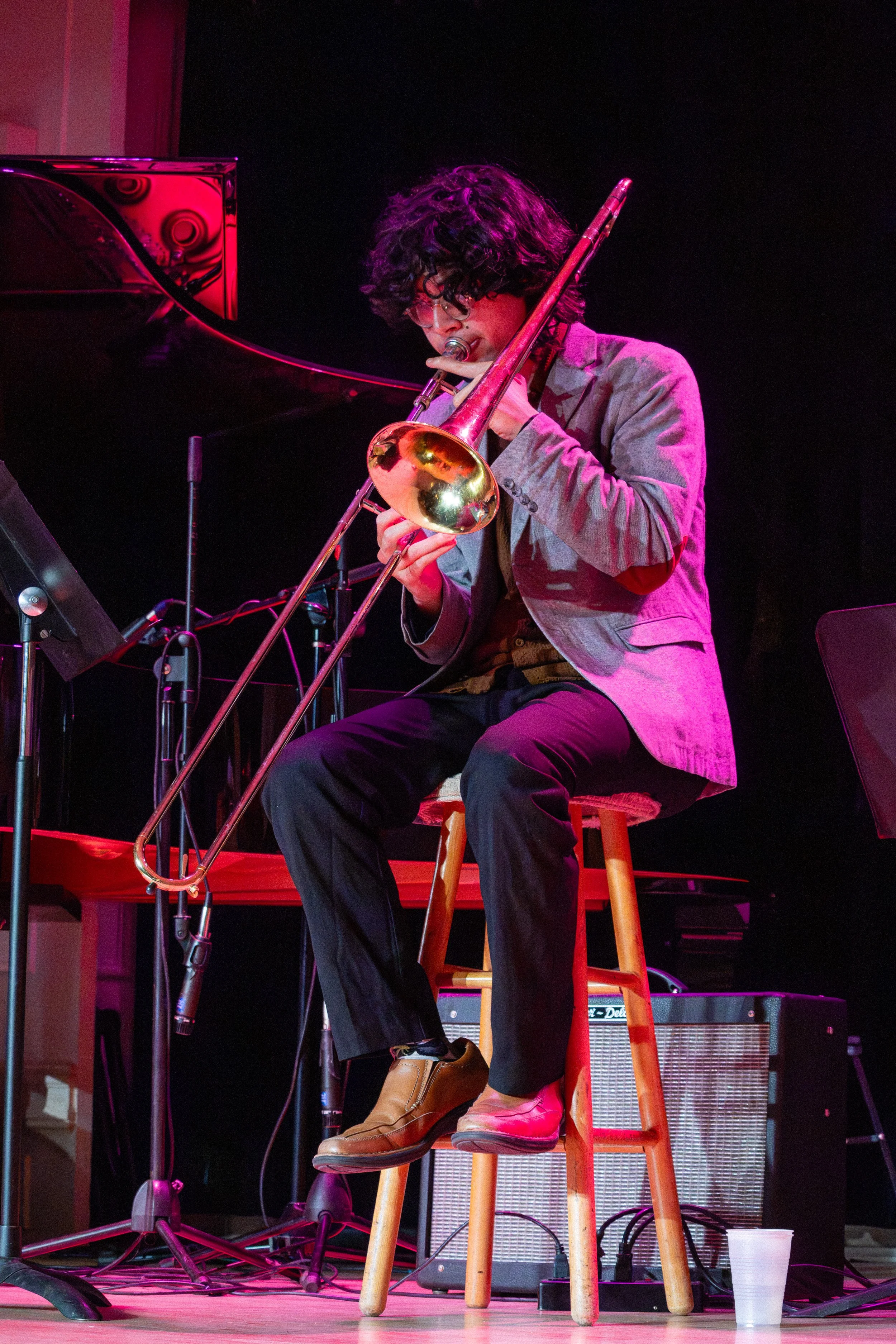A musician with curly hair, glasses, wearing a gray blazer and brown shoes, is playing the trombone while seated on a wooden stool on stage with pink and purple lighting, with musical equipment and a plastic cup nearby.