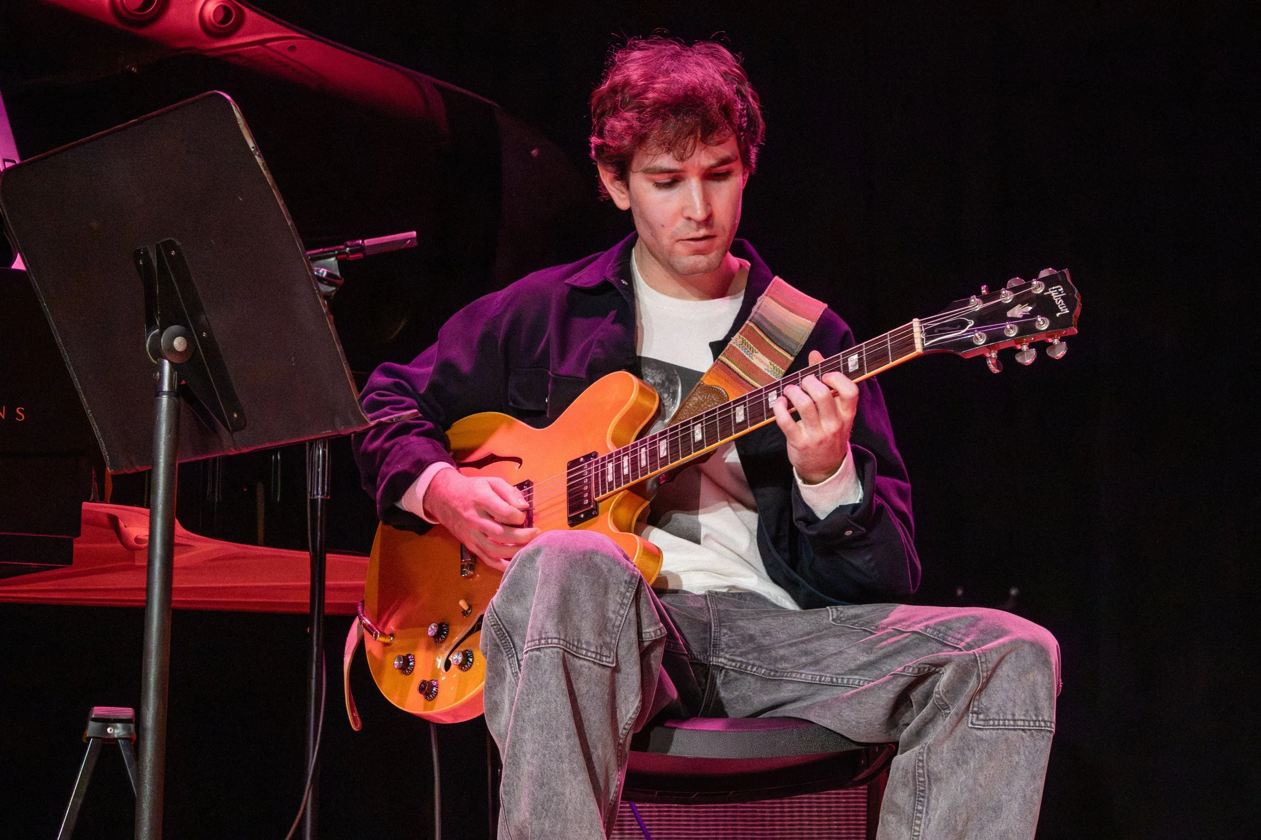 A young man playing an electric guitar on stage with a black background, wearing a dark jacket over a graphic t-shirt and jeans, surrounded by musical equipment.