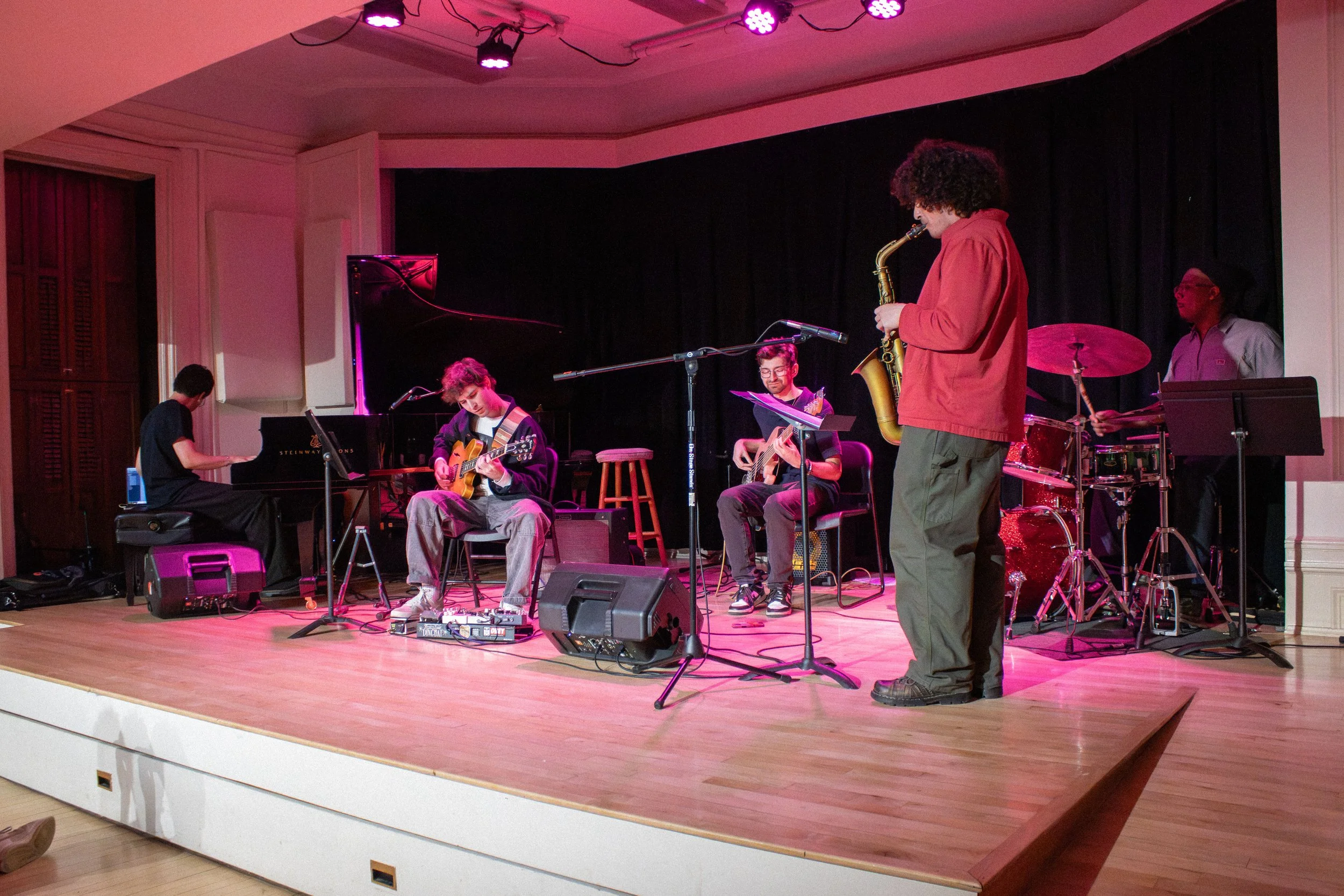 A jazz band performing on stage, featuring musicians playing piano, guitar, saxophone, and drums, with a black curtain backdrop and stage lighting.