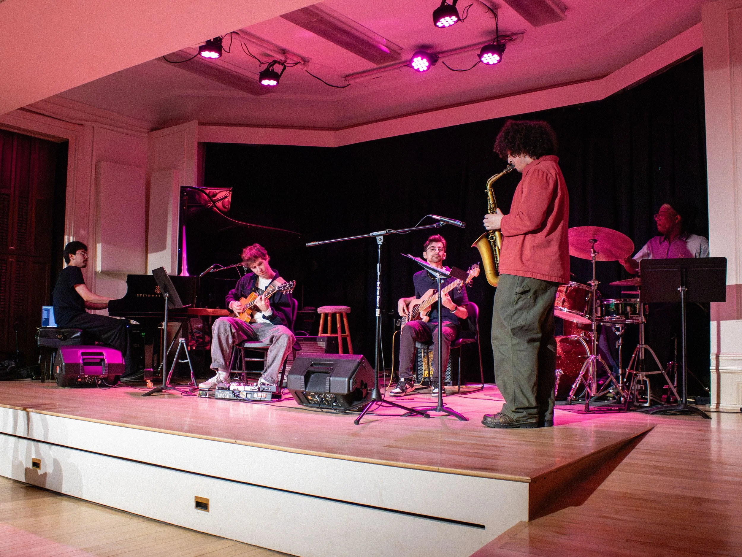 A group of musicians performing on a stage with a black curtain backdrop, including a pianist, guitarist, drummer, and a saxophonist in a red jacket, with purple stage lighting.