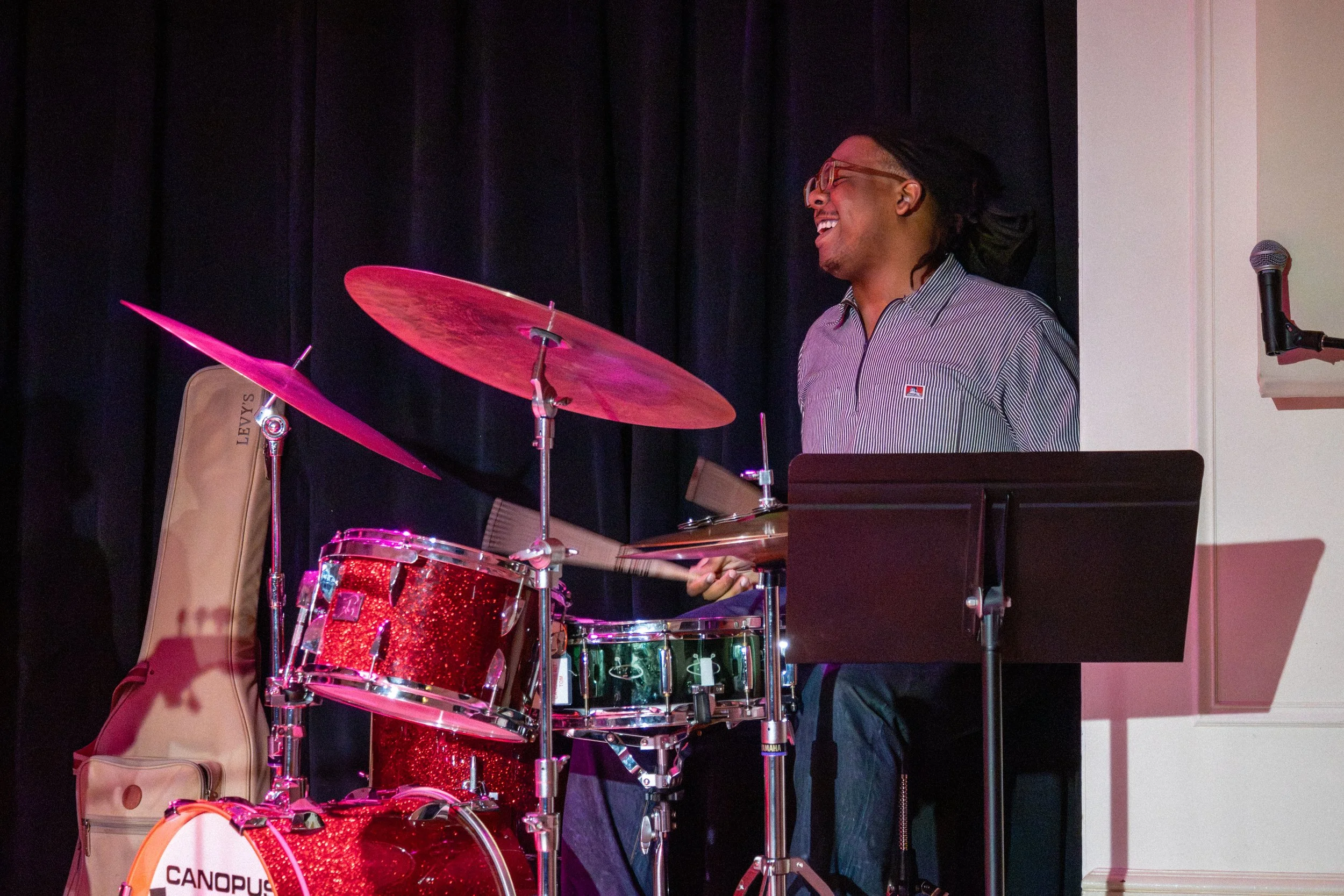 A man with glasses and dreadlocks playing a red drum set on stage, smiling and laughing, with a black curtain background and a microphone on a stand nearby.