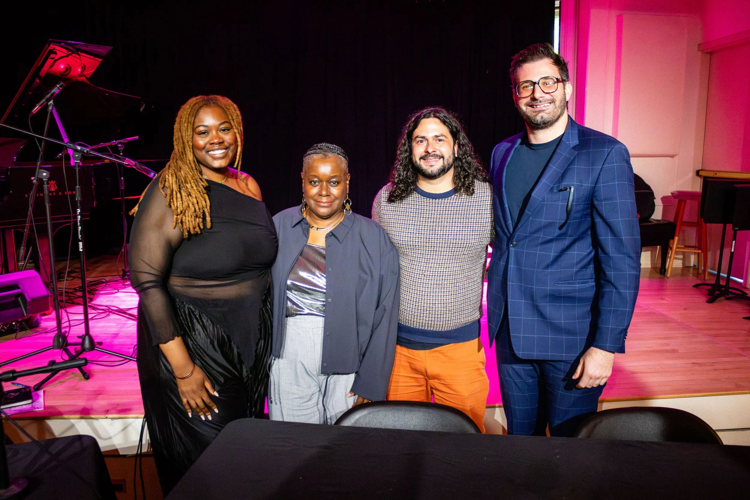 A group of five diverse people standing together in a music venue or studio with a piano and microphones in the background. They are smiling and facing the camera.
