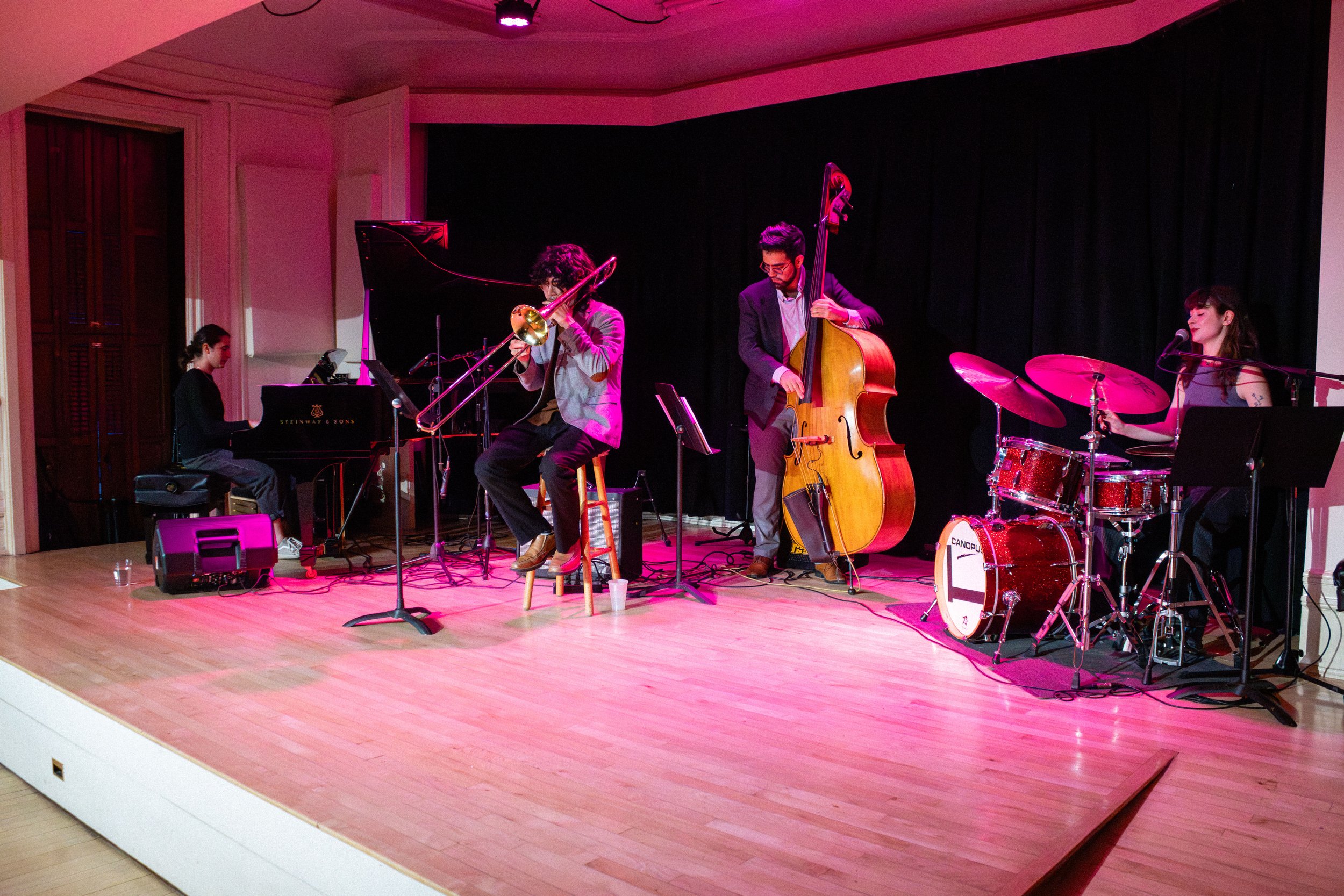 A jazz band performing on a stage with four musicians: a pianist, a trombone player, a double bass player, and a drummer, against a black curtain backdrop, with pink stage lighting.