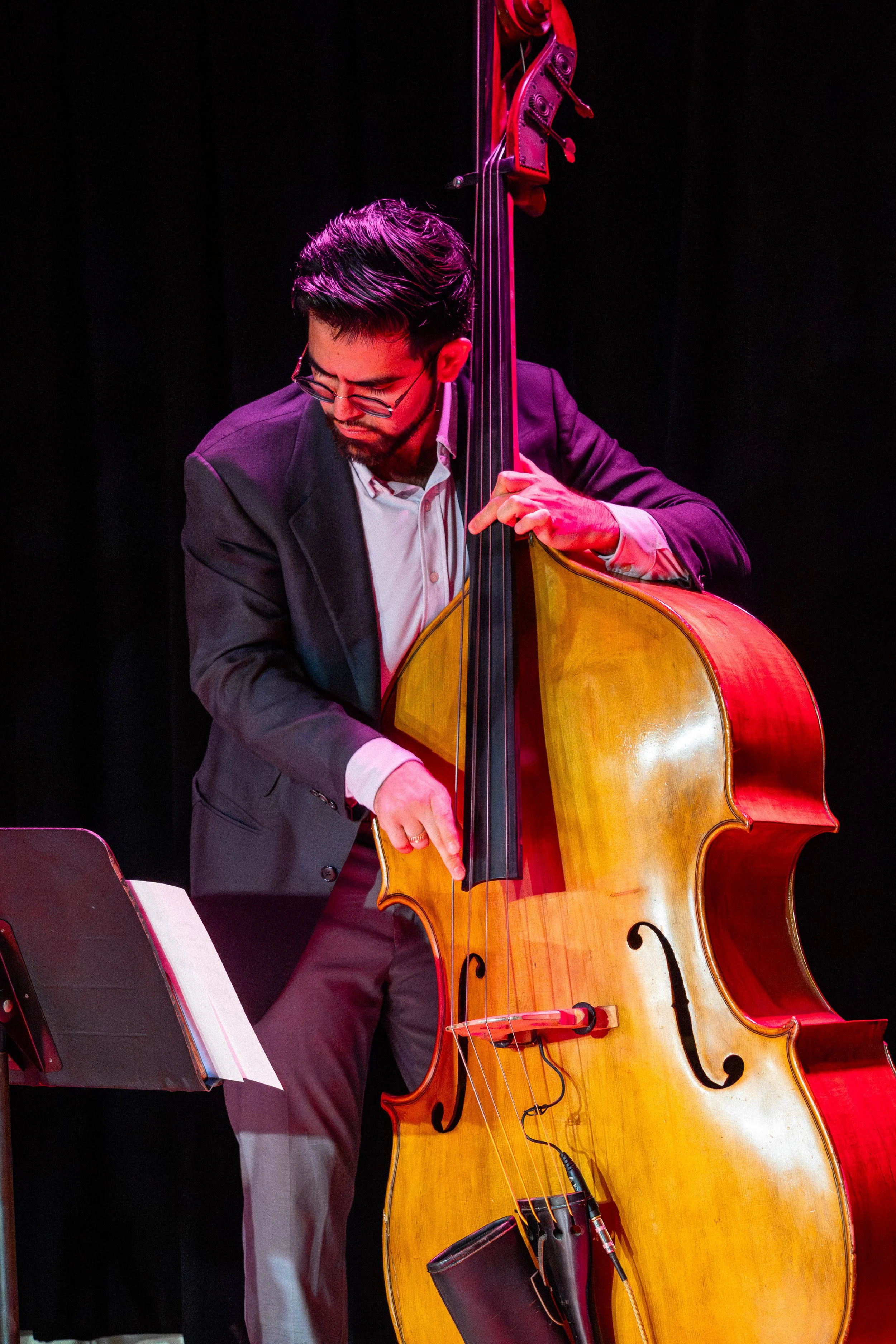 A man in a suit playing a double bass on stage with black background.