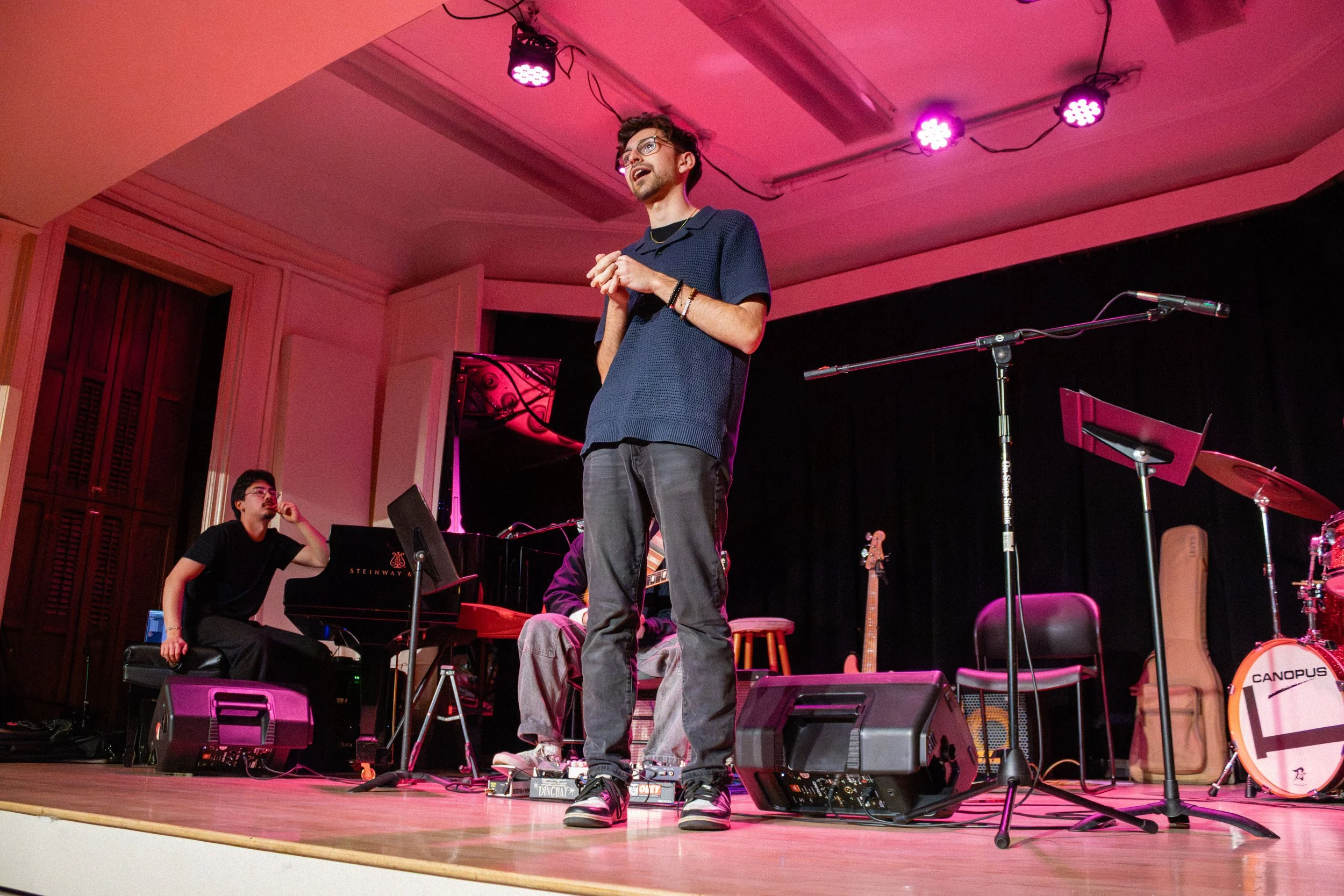 A man stands on a stage with pink lighting, singing into a microphone. Two other musicians are seated nearby, one at a piano and the other with a guitar. The stage has musical instruments and equipment, including a drum and a music stand, and the bac