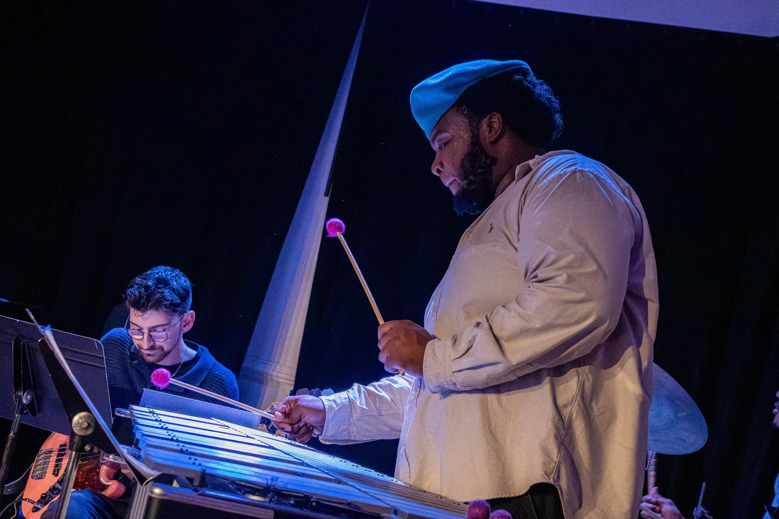 A man in a beige shirt and blue cap playing percussion with a drumstick at a musical performance, with a younger man in glasses also playing an instrument nearby, set against a dark background.