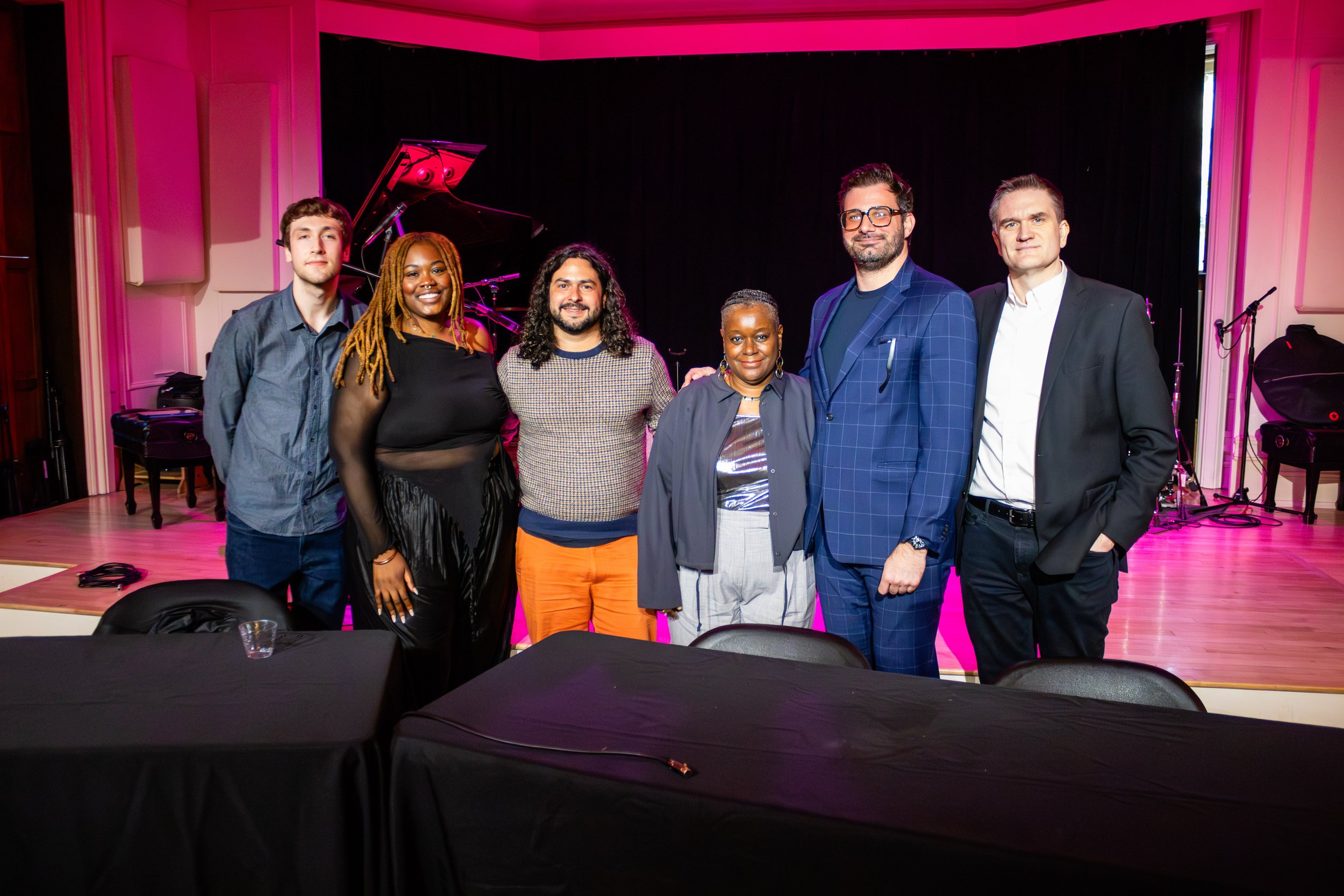 Group of seven diverse people standing together on a stage with black curtains, pink lighting, and musical instruments in the background, smiling and posing for a photo.
