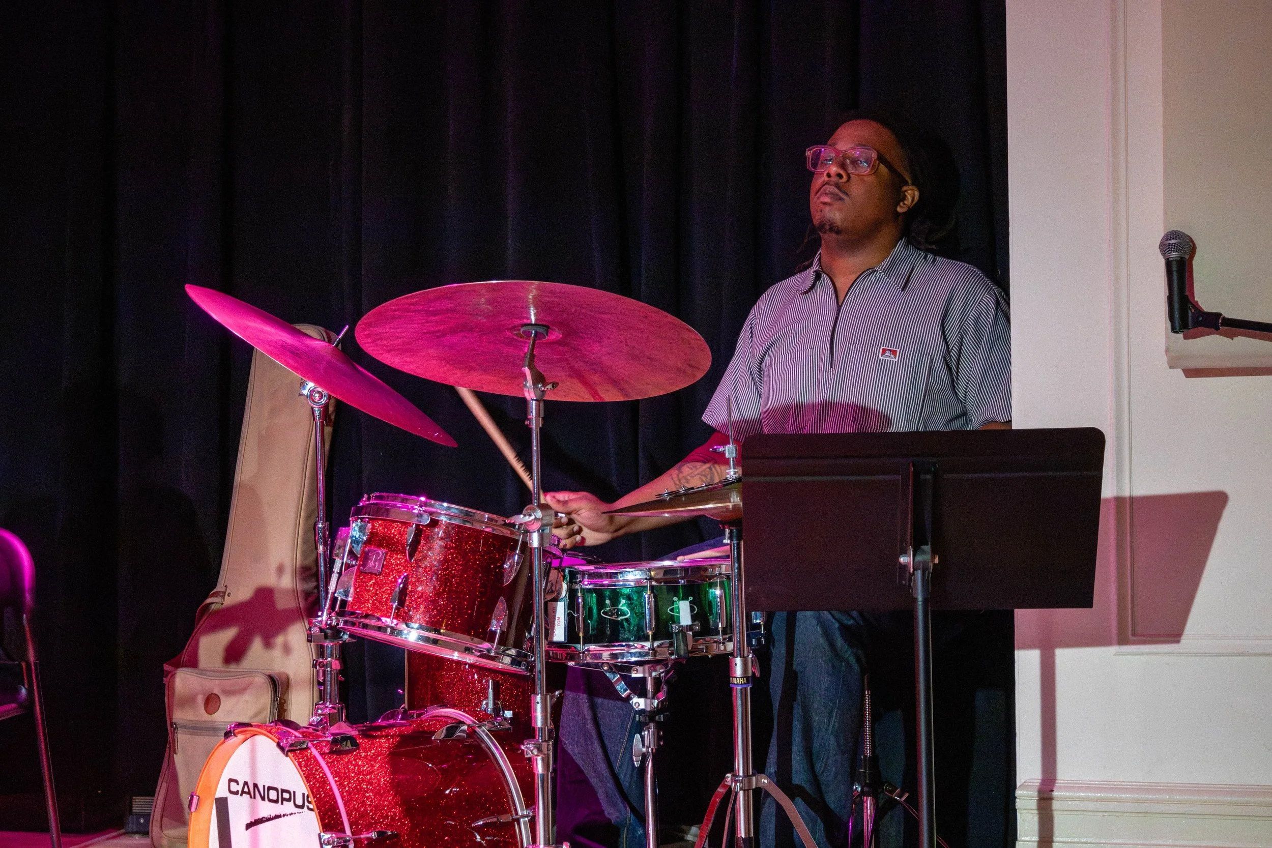 A man wearing glasses and a striped shirt playing a red drum set on a stage with a dark curtain background and a microphone nearby.