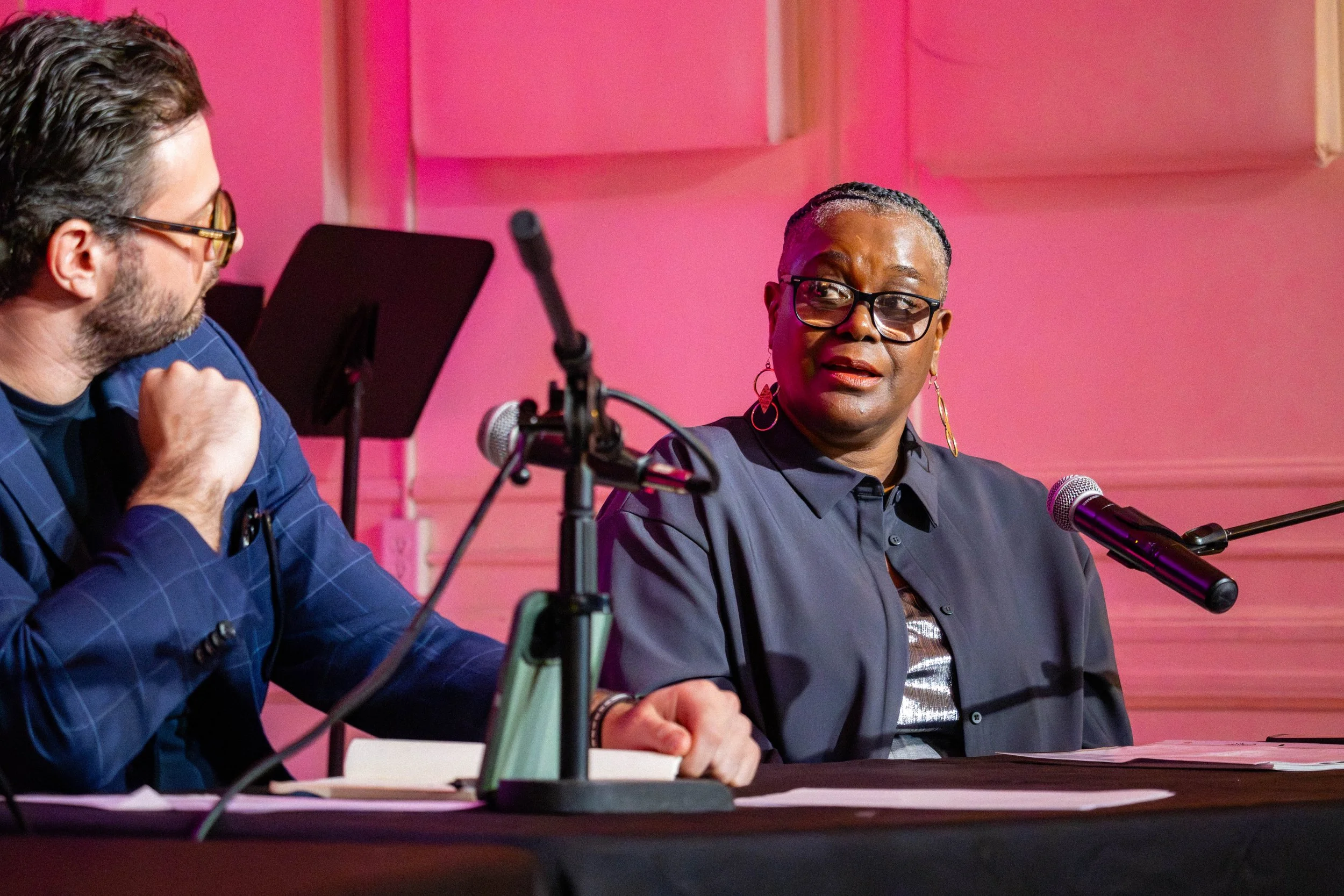 A man and a woman sitting at a table during a discussion or interview, with microphones nearby, against a pink background.