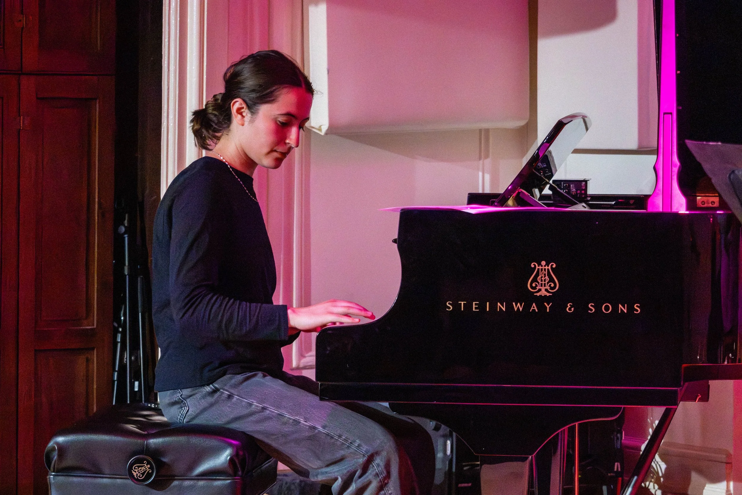 Young woman with short dark hair playing a black Steinway & Sons grand piano, sitting on a black piano bench in a room with wooden door and pink lighting.