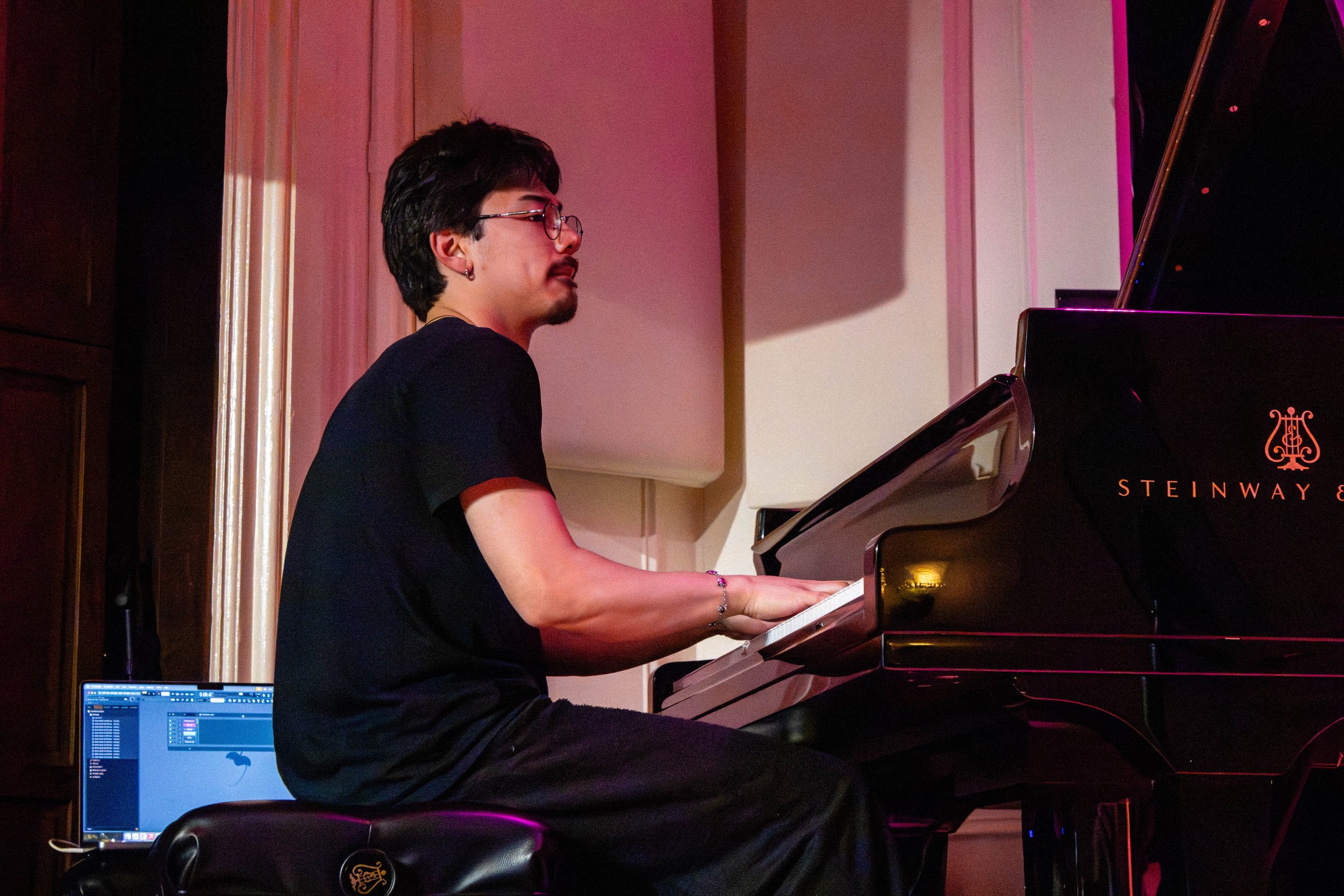 A person with dark hair, glasses, earrings, and a goatee playing a Steinway grand piano in a room with pink lighting.