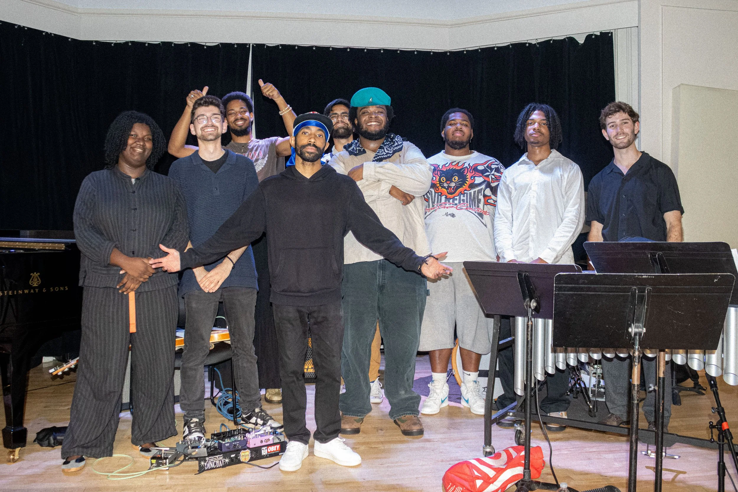 Group of nine people standing together on a stage, smiling, with musical instruments and equipment in front of them, black curtains in the background.