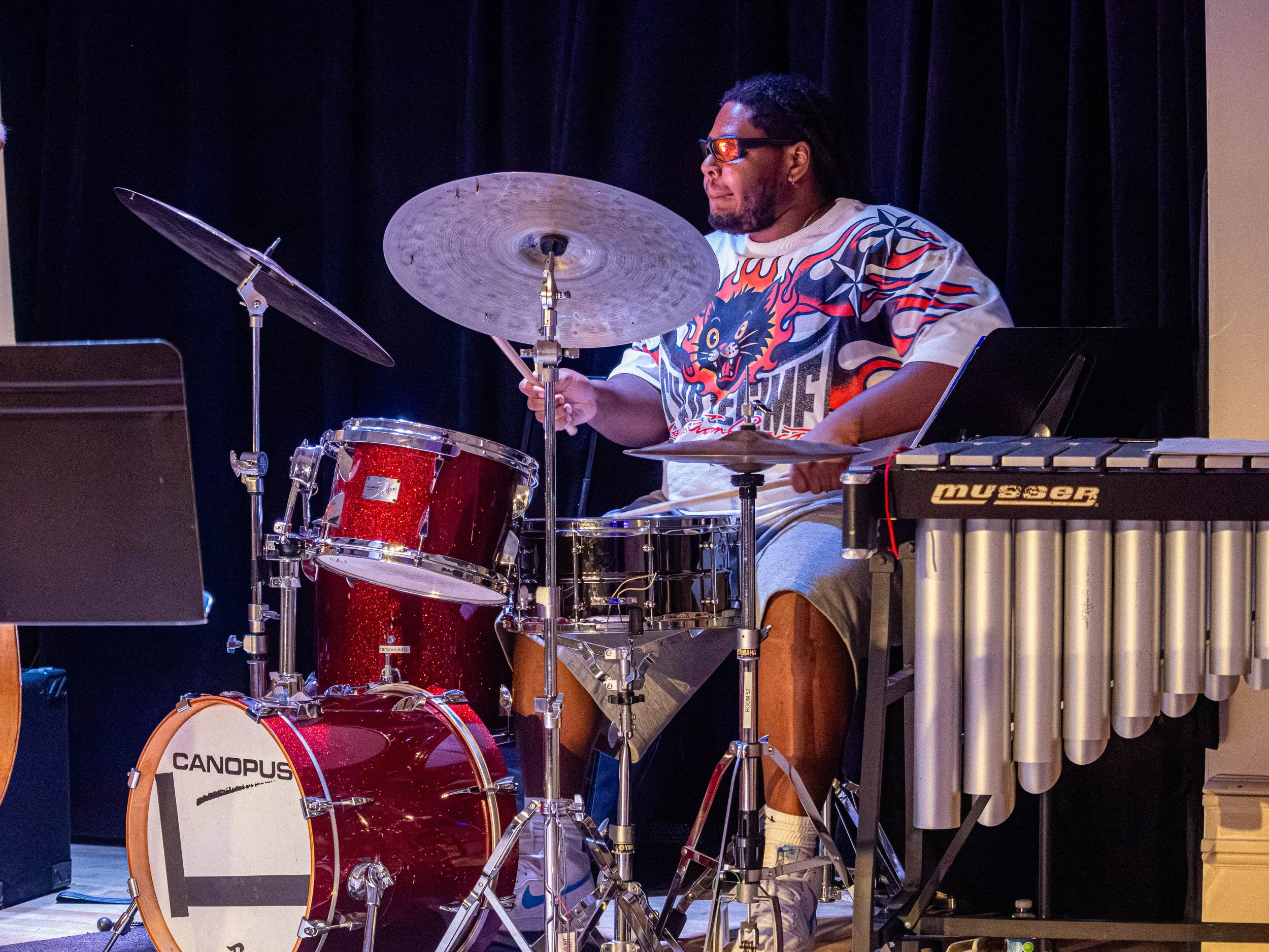 A musician playing a drum set on stage, wearing sunglasses and a colorful shirt with a lion graphic.