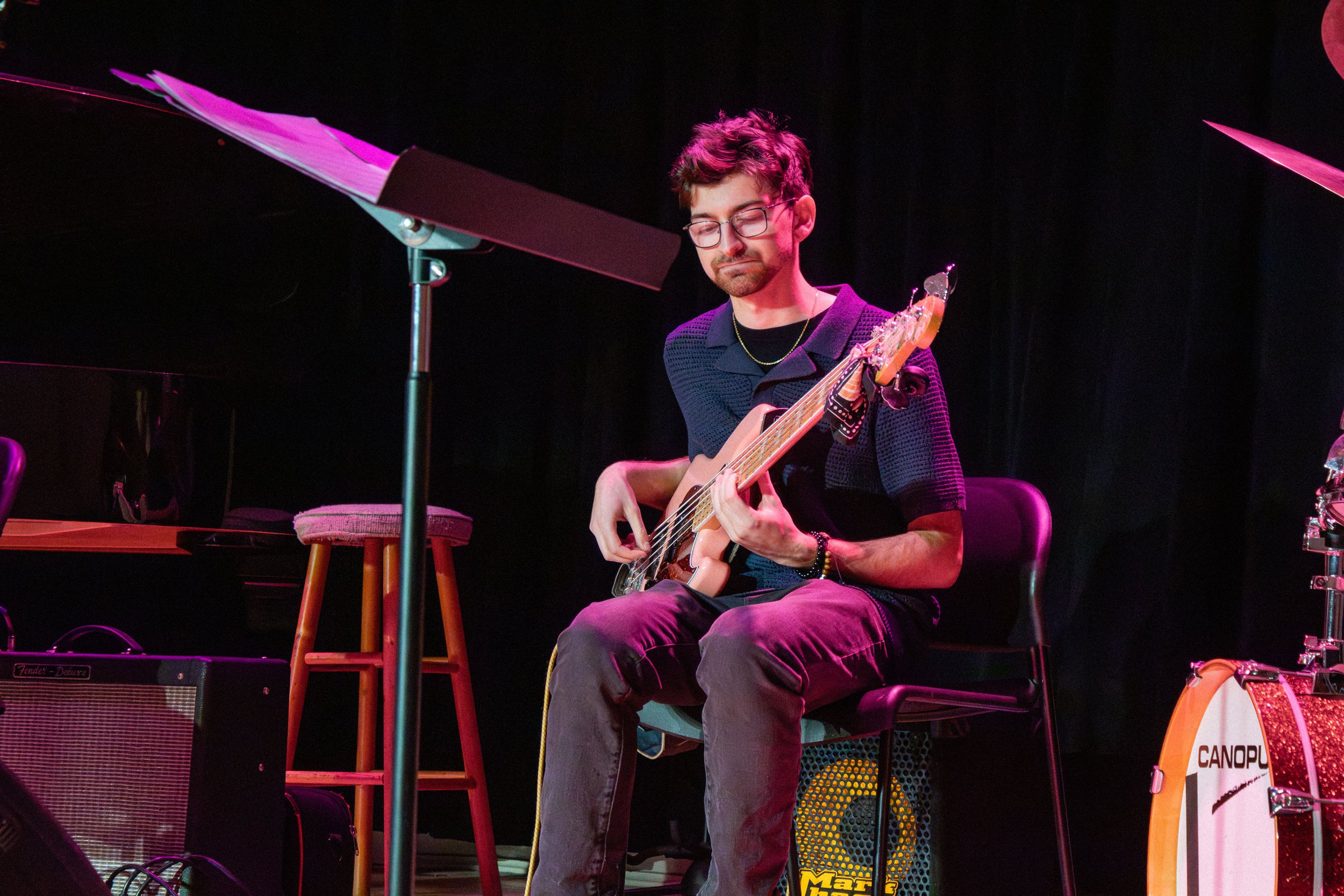 A young man with glasses, wearing a black shirt, playing an electric guitar on stage. There are musical equipment and furniture around him, with stage lighting creating a vibrant atmosphere.