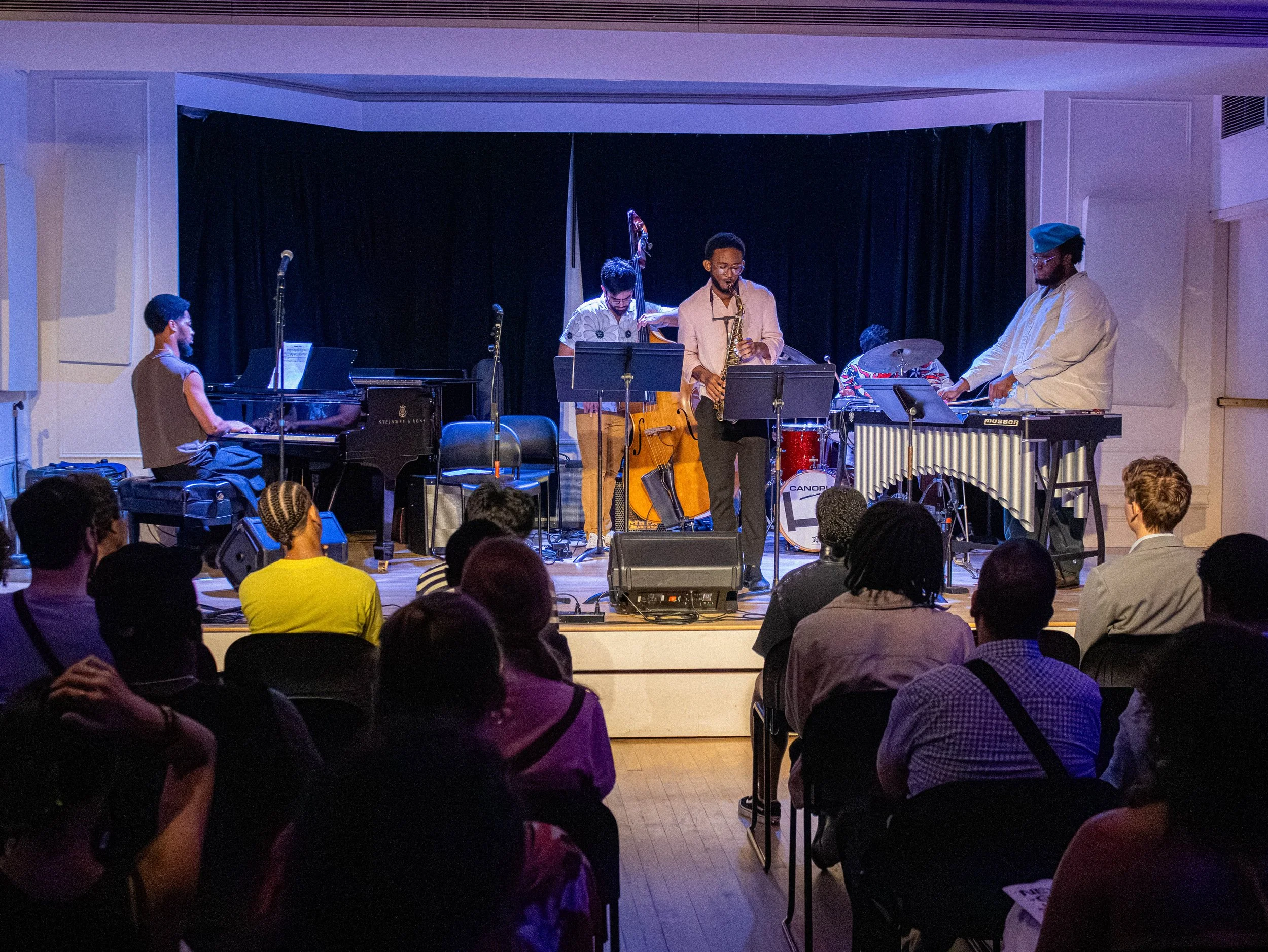 A jazz band performing on stage with musicians playing piano, upright bass, drums, saxophone, and vibraphone, in front of an audience.