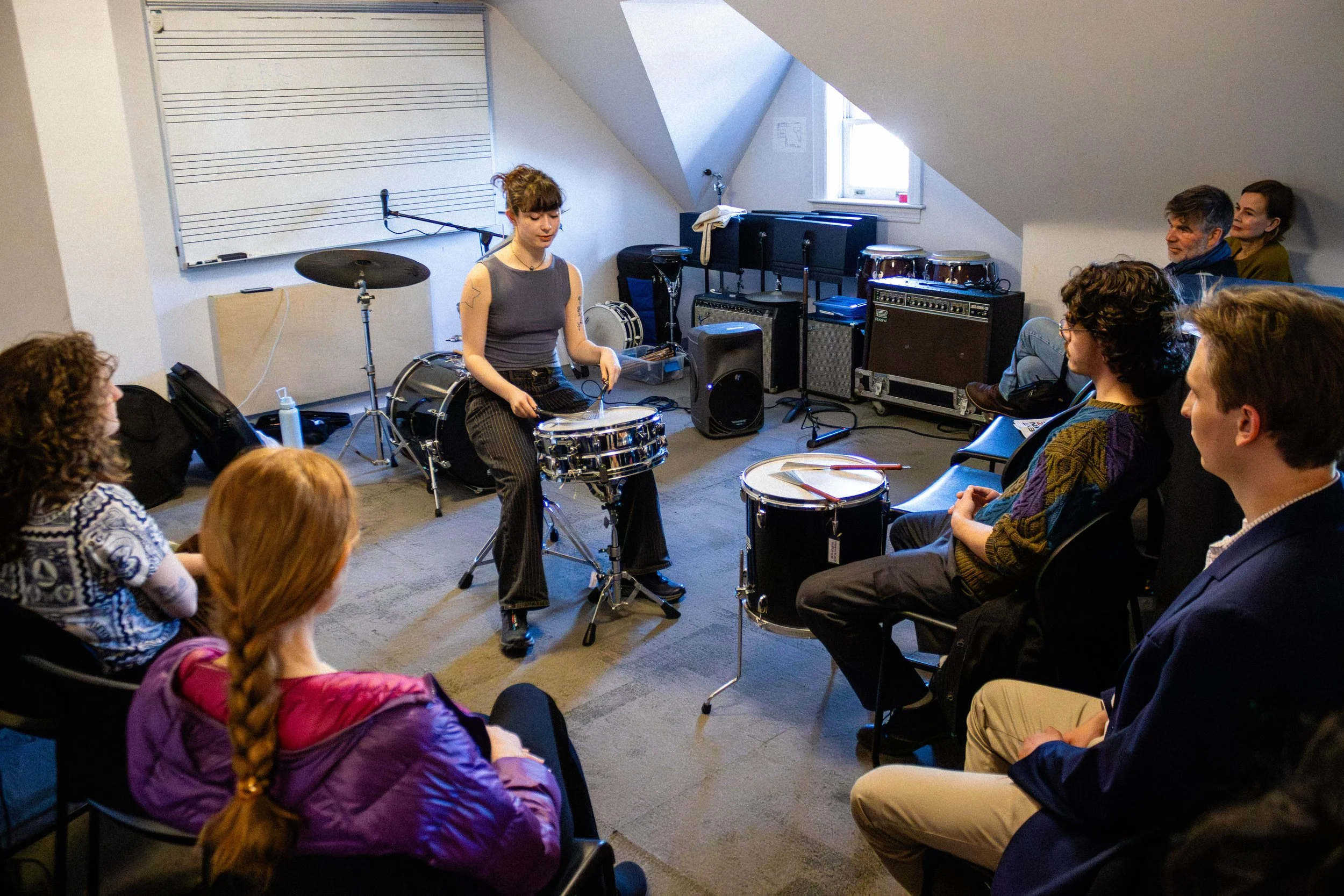 A woman demonstrates playing drums to a seated audience in a room with musical equipment and a music whiteboard on the wall.