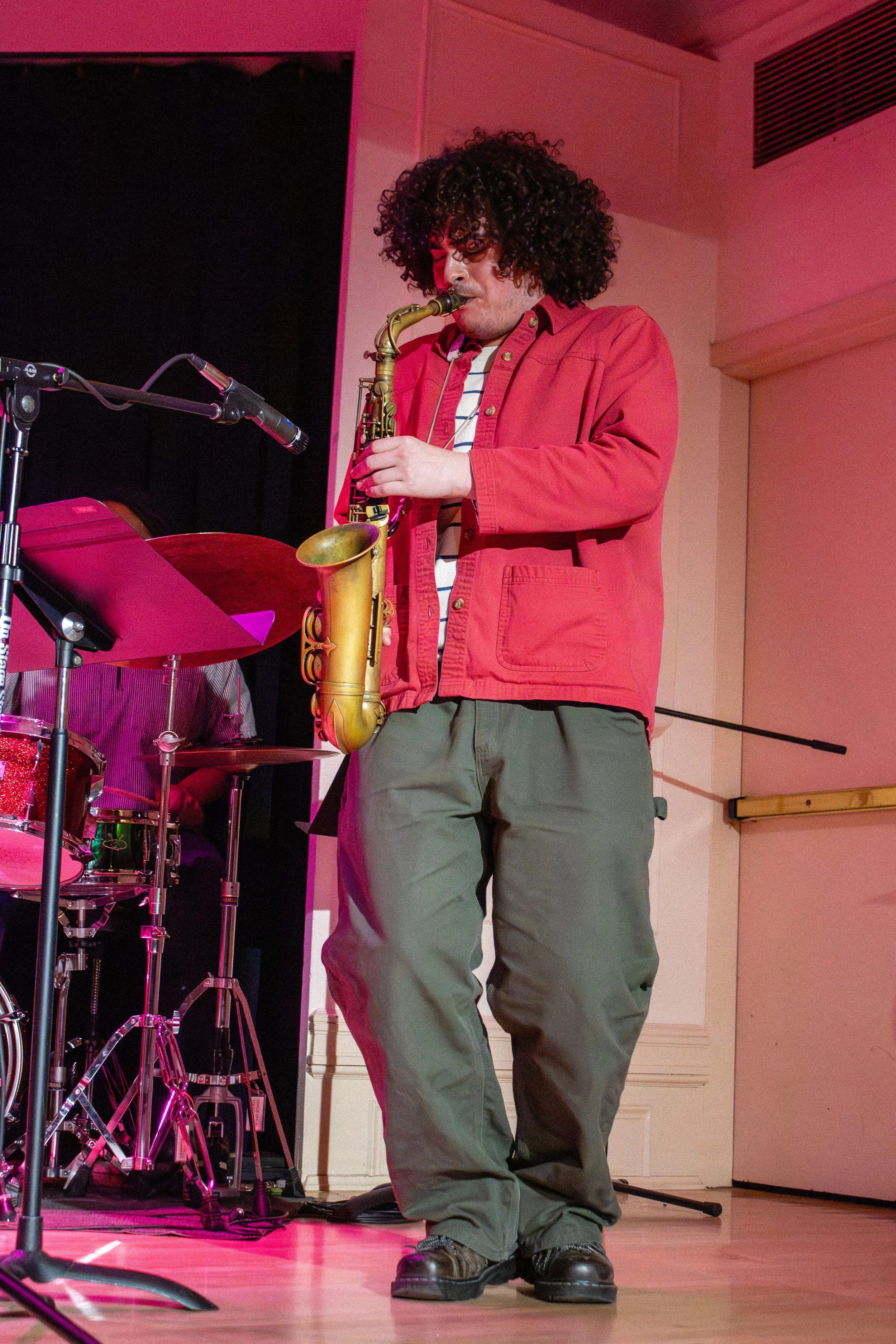 A man with curly dark hair playing a saxophone during a live performance. He is wearing a red jacket over a striped shirt and green pants, standing on a wooden stage with musical equipment around him, illuminated by pink stage lighting.