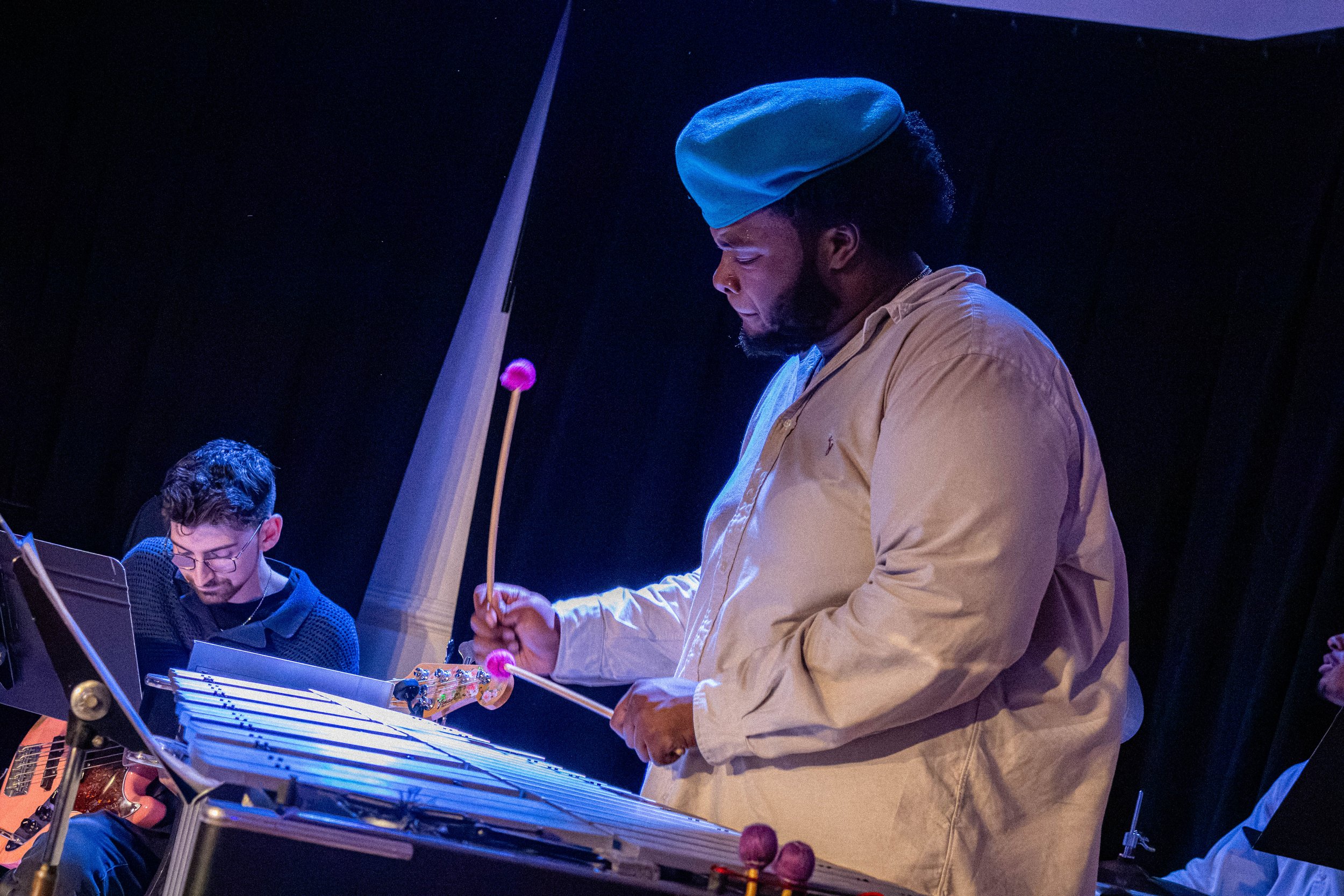 A musician playing a vibraphone during a performance, with another musician reading sheet music nearby, in a dark setting.