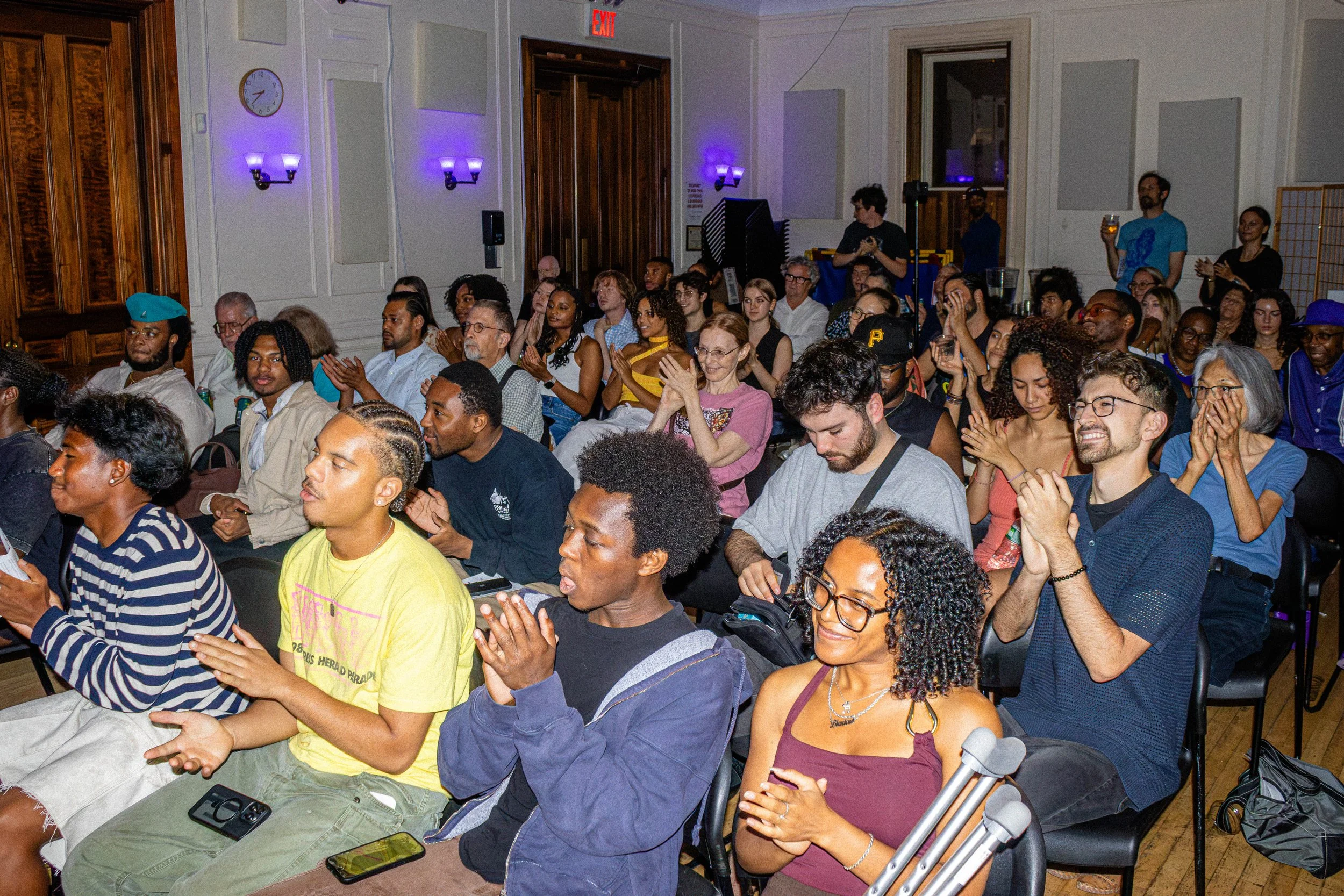 A diverse group of people sitting and clapping at an indoor event, with some using smartphones and others smiling and engaging, in a room with white walls and purple lighting.