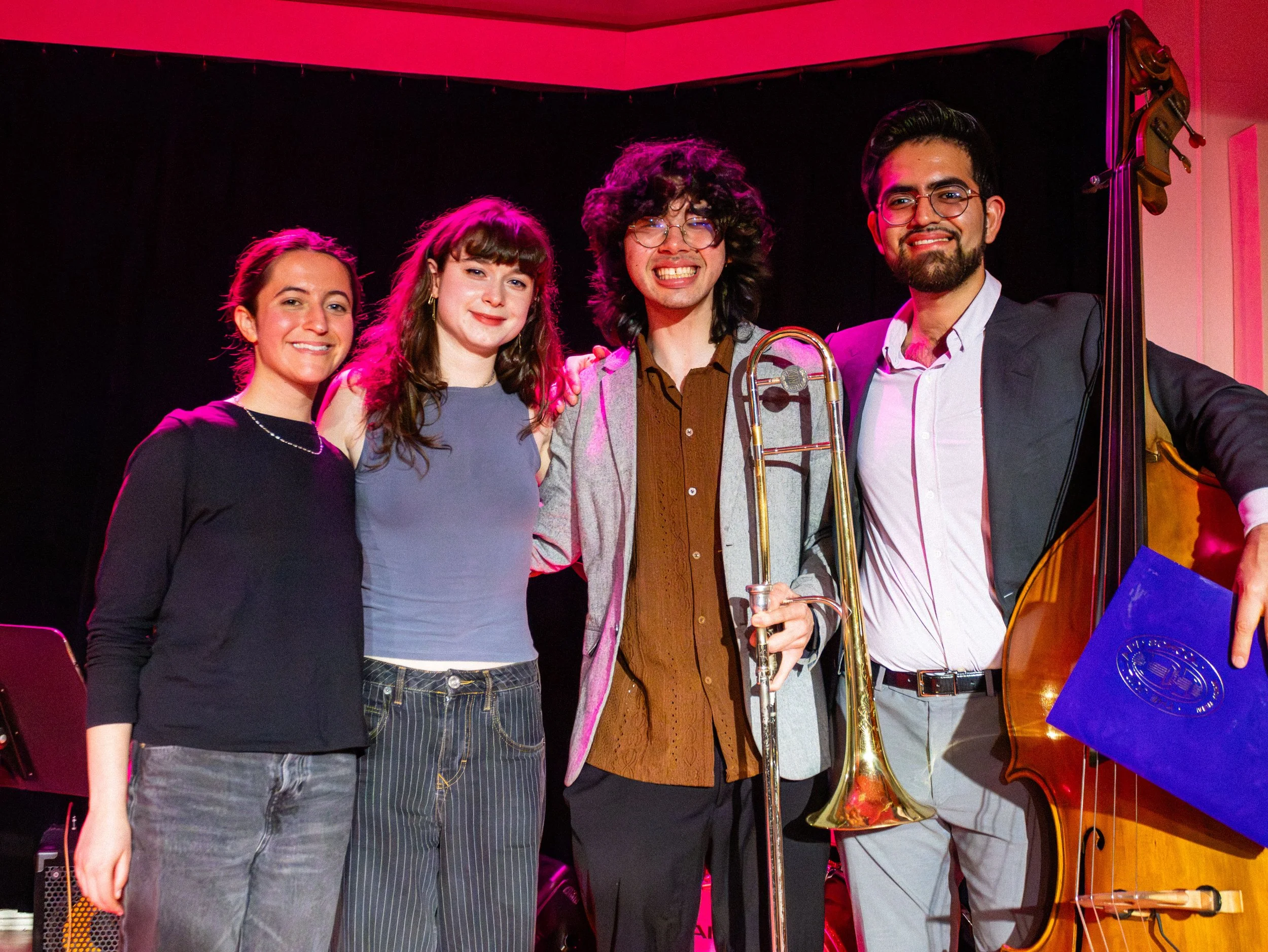 Group of four young adults smiling on stage, with musical instruments including a trombone and upright bass, against a black backdrop with pink lighting.