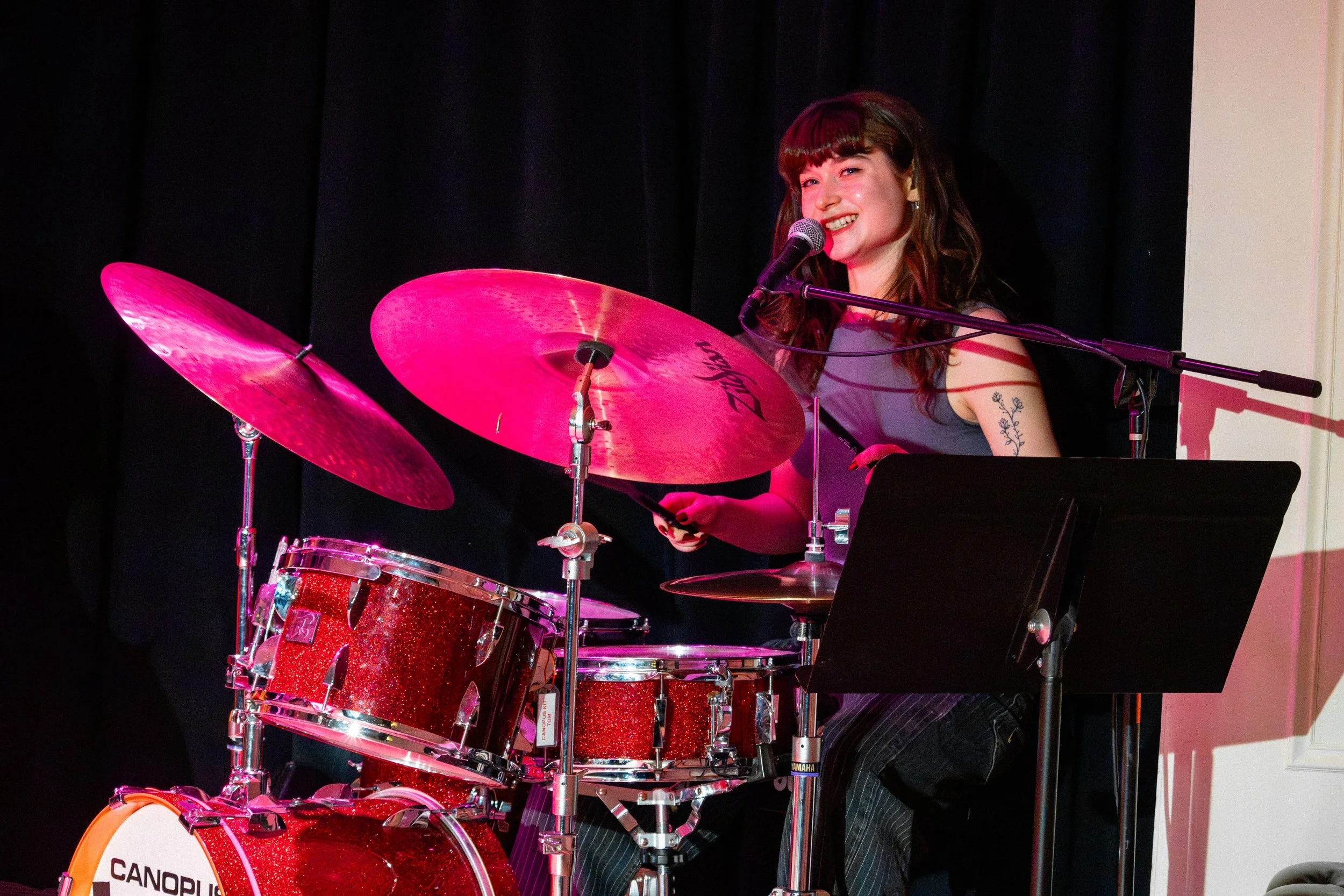 A young woman with brown hair and bangs, smiling and singing into a microphone, sits behind a red glittery drum set and plays drums on stage with a black curtain background.