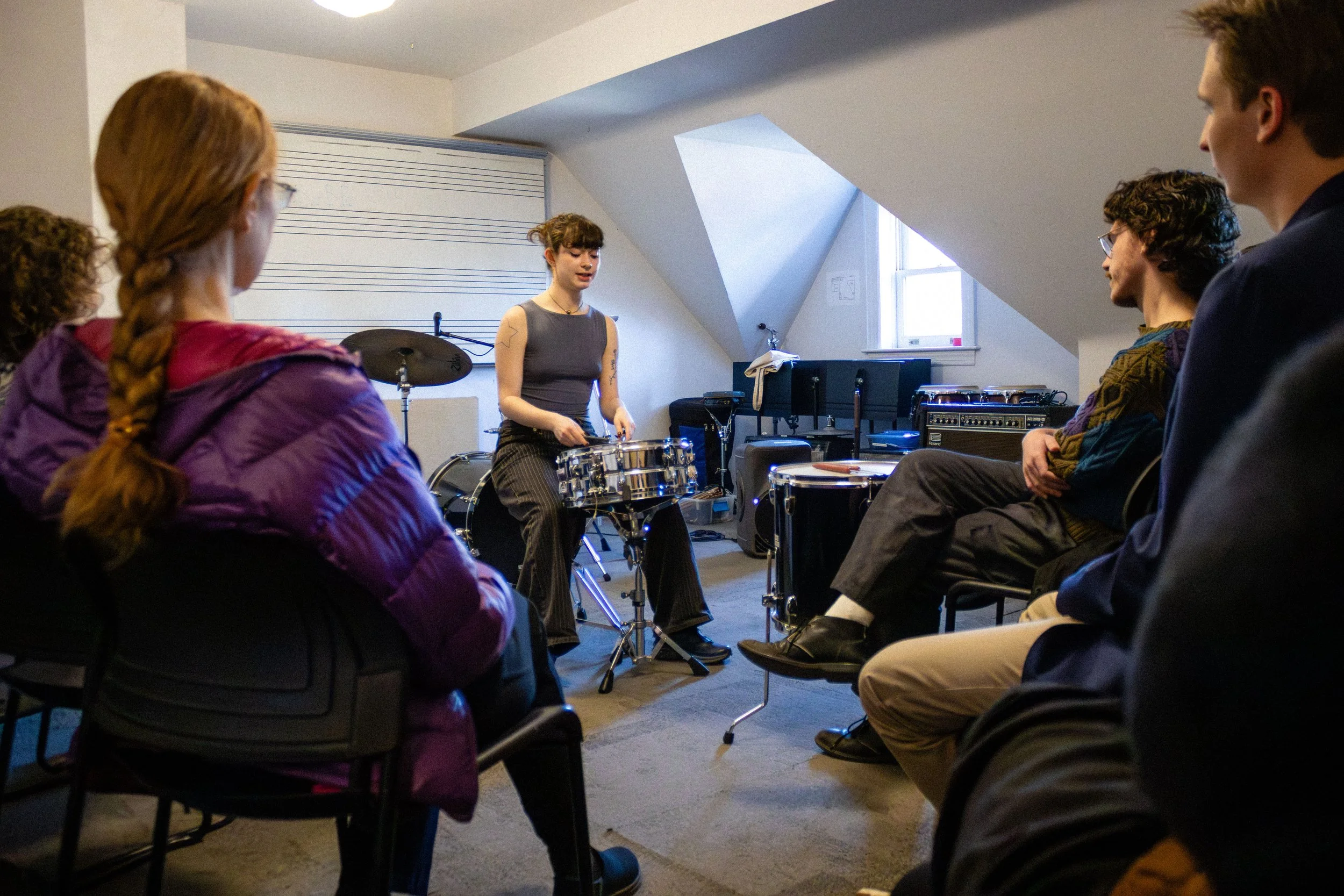 A woman demonstrating drums to a group of people seated in a circle in a room with sloped ceilings and a small window.