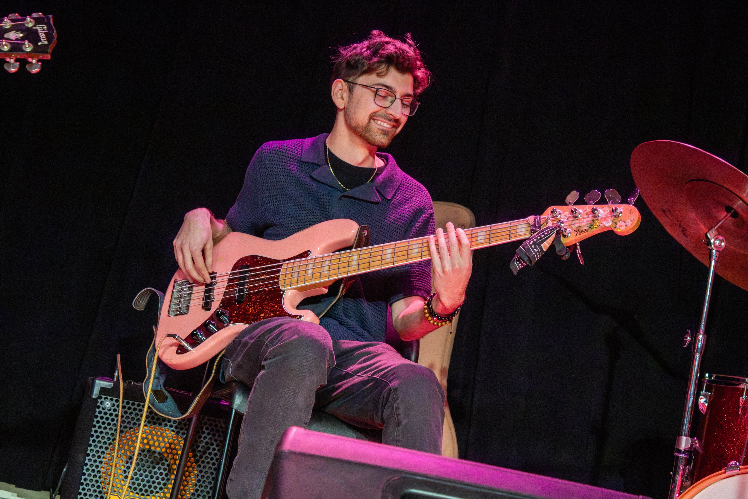 A young man with glasses and a beard playing an electric bass guitar on stage, smiling, with a drum set and a black background behind him.