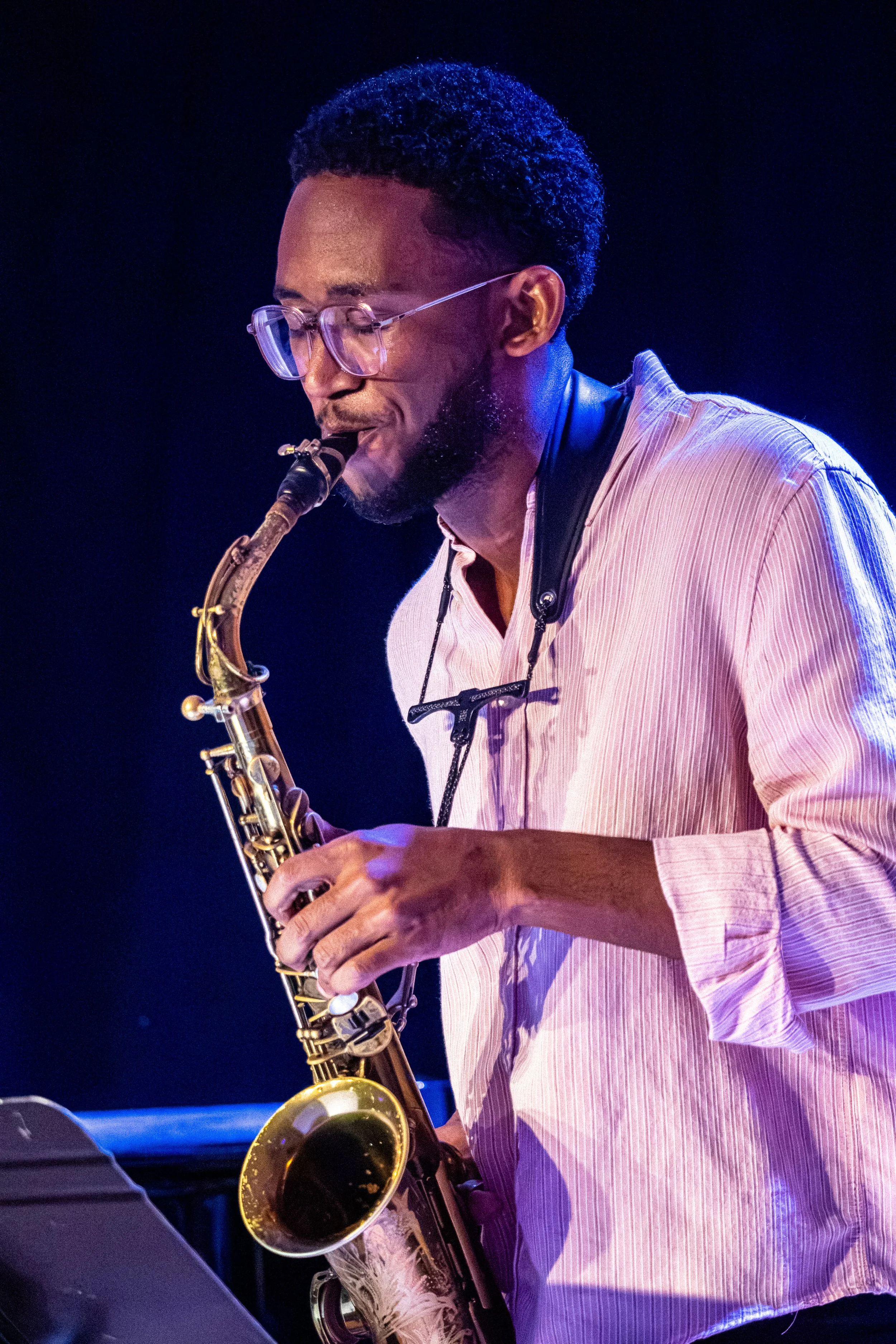 A man with glasses and a beard playing a saxophone on stage, wearing a light pink and white striped shirt, with purple stage lighting in the background.
