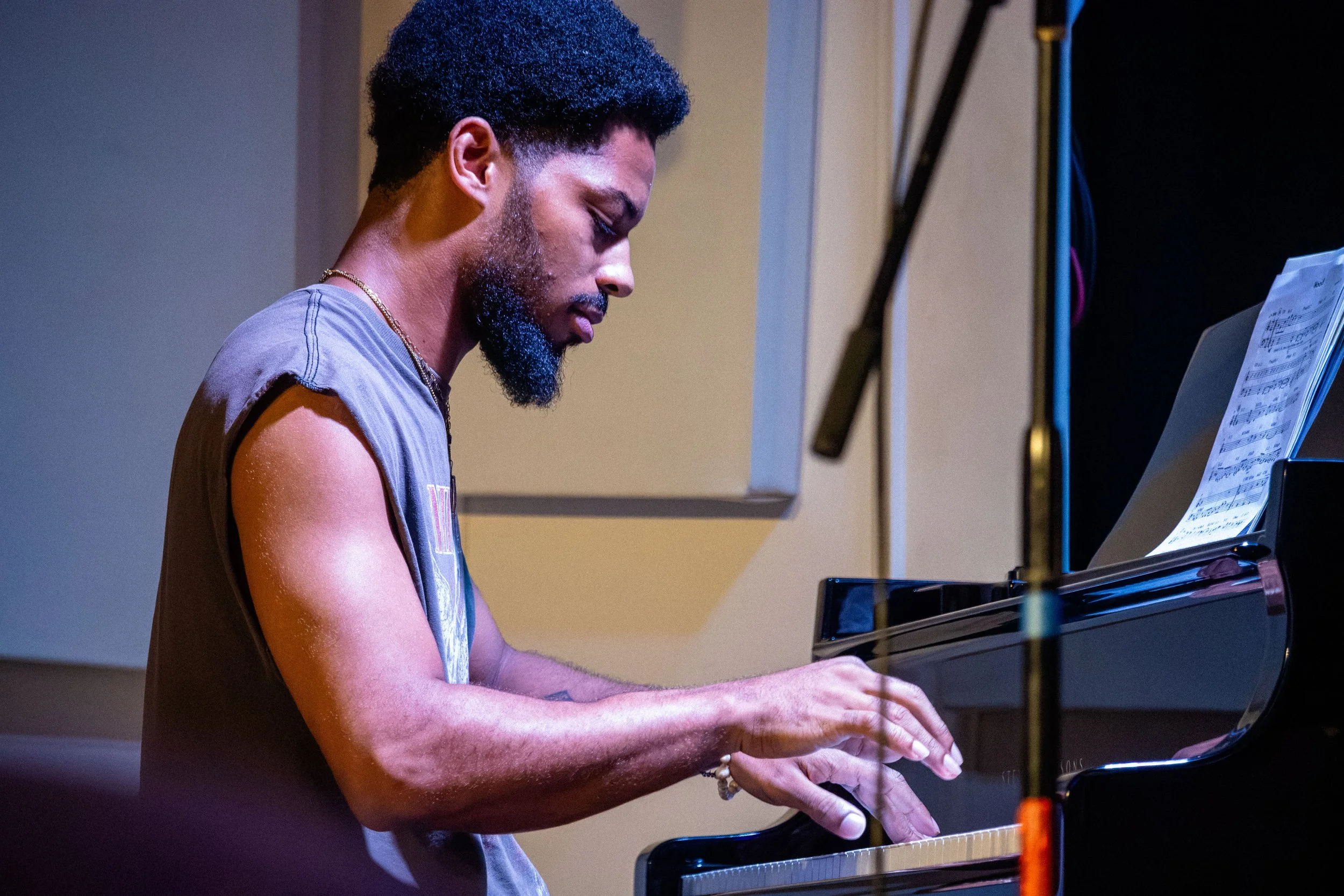 A young man with a beard and curly hair playing the piano, wearing a sleeveless shirt and a gold necklace.