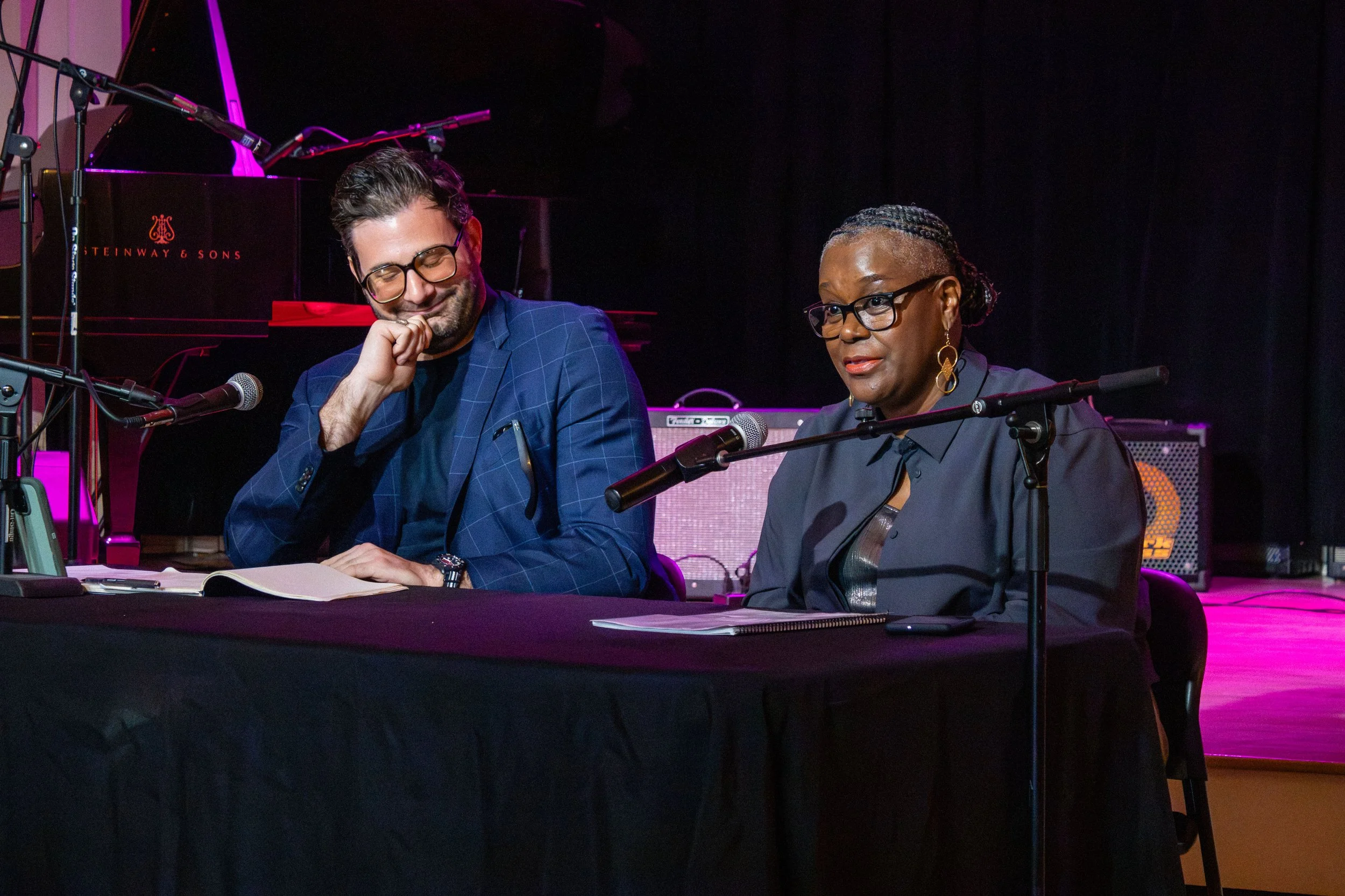 Two people, a man and a woman, sitting at a table with microphones, books, and papers, in a music studio or performance space with a grand piano in the background and stage lighting.